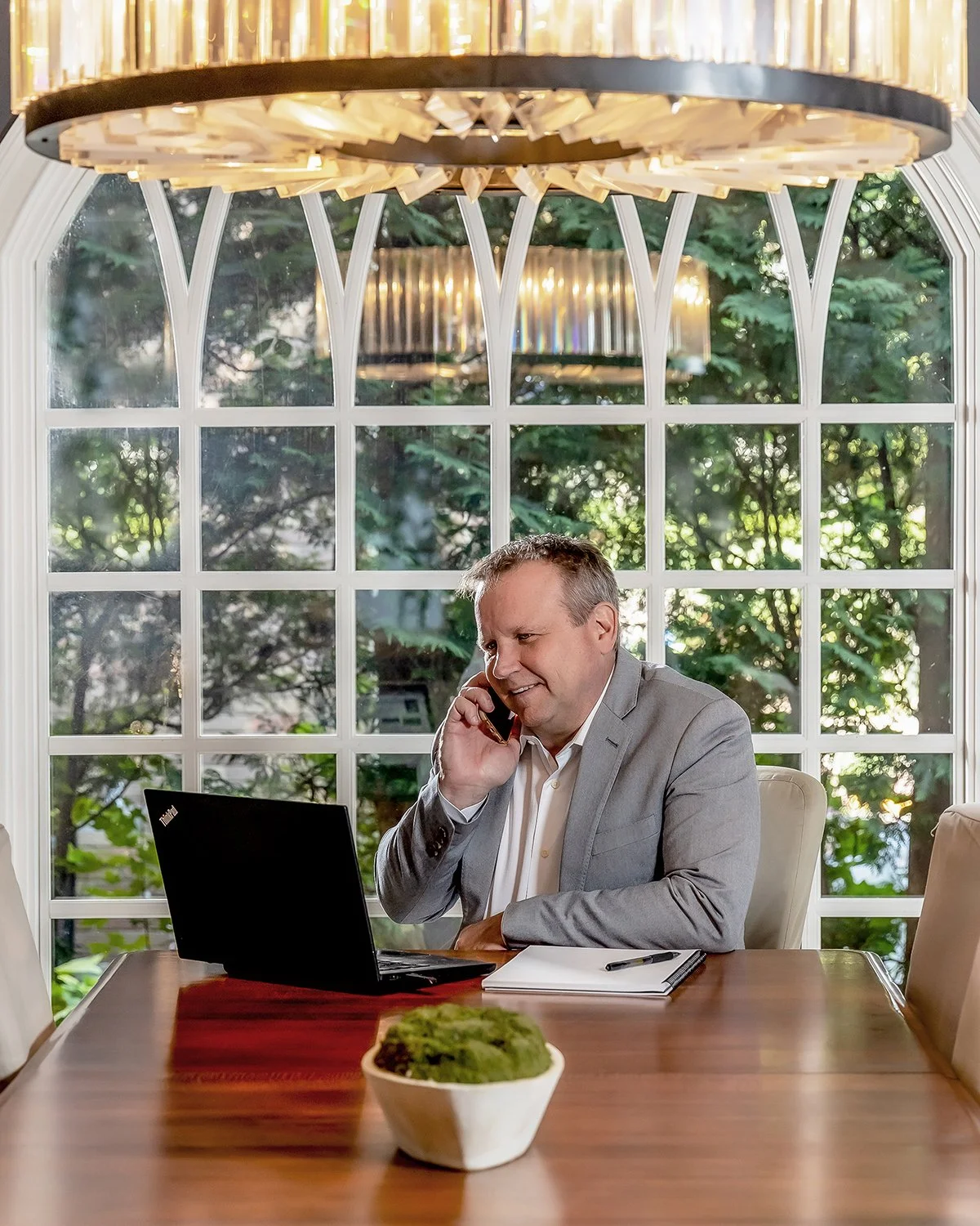 Smiling Male Executive in a Grey Suit at a Wooden Table Working on a Laptop and Talking on the Phone in Front of a Large Window