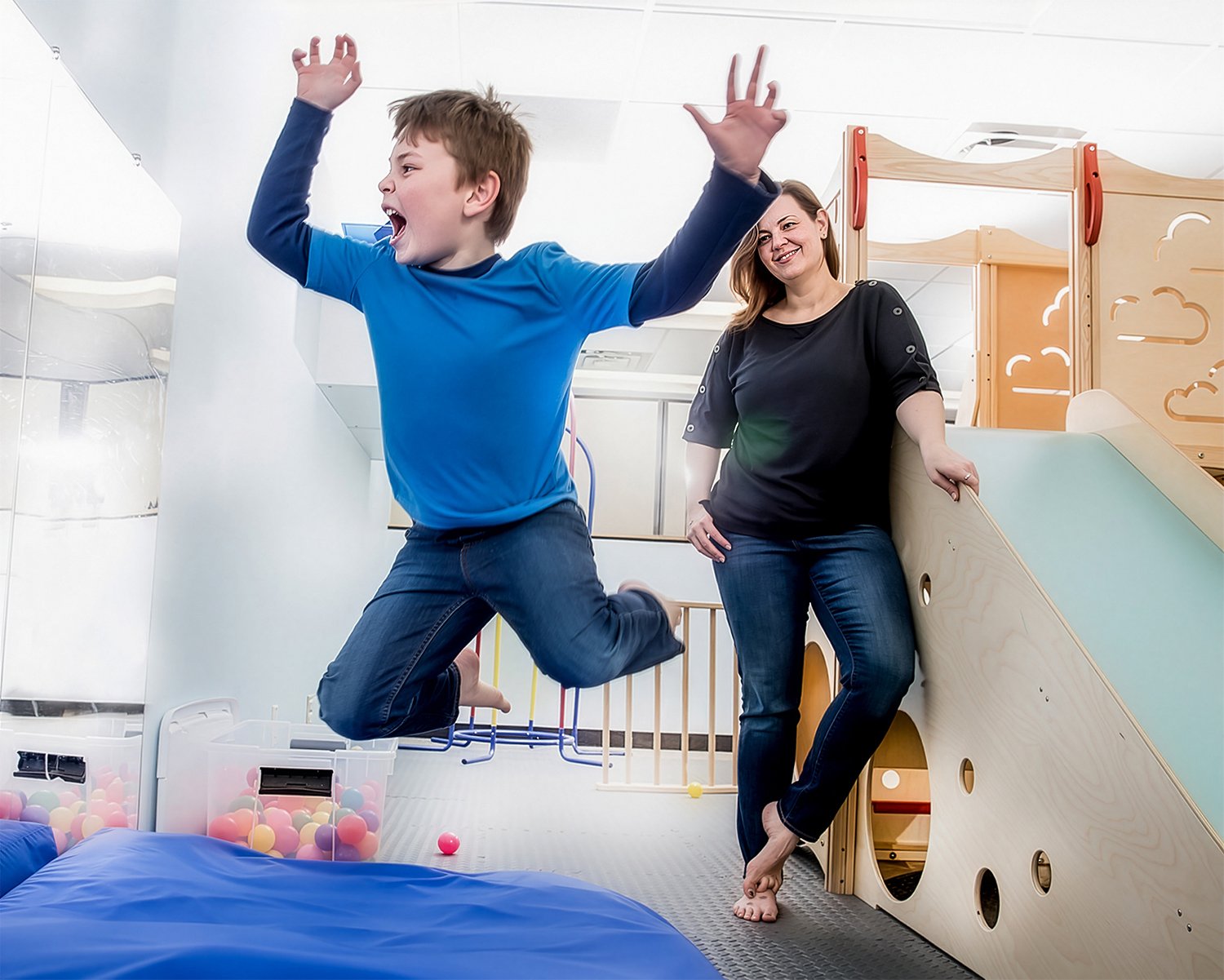 Autistic Boy Running and Jumping in Play Area with Mother Looking On.  Healthcare Lifestyle Photography