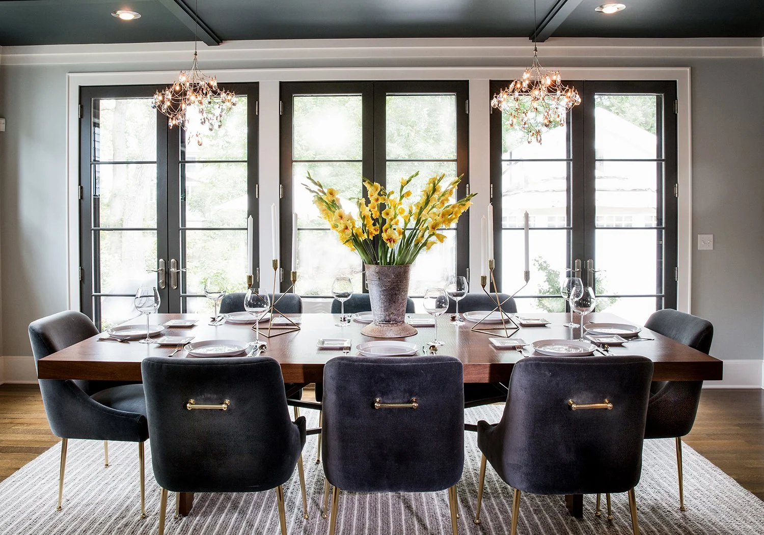 Luxury Dining Room Interior with a Wood Table, Dark Velvet Chairs, and a Large Yellow Flower Arrangement Featuring Large Glass French Doors and Twin Chandeliers