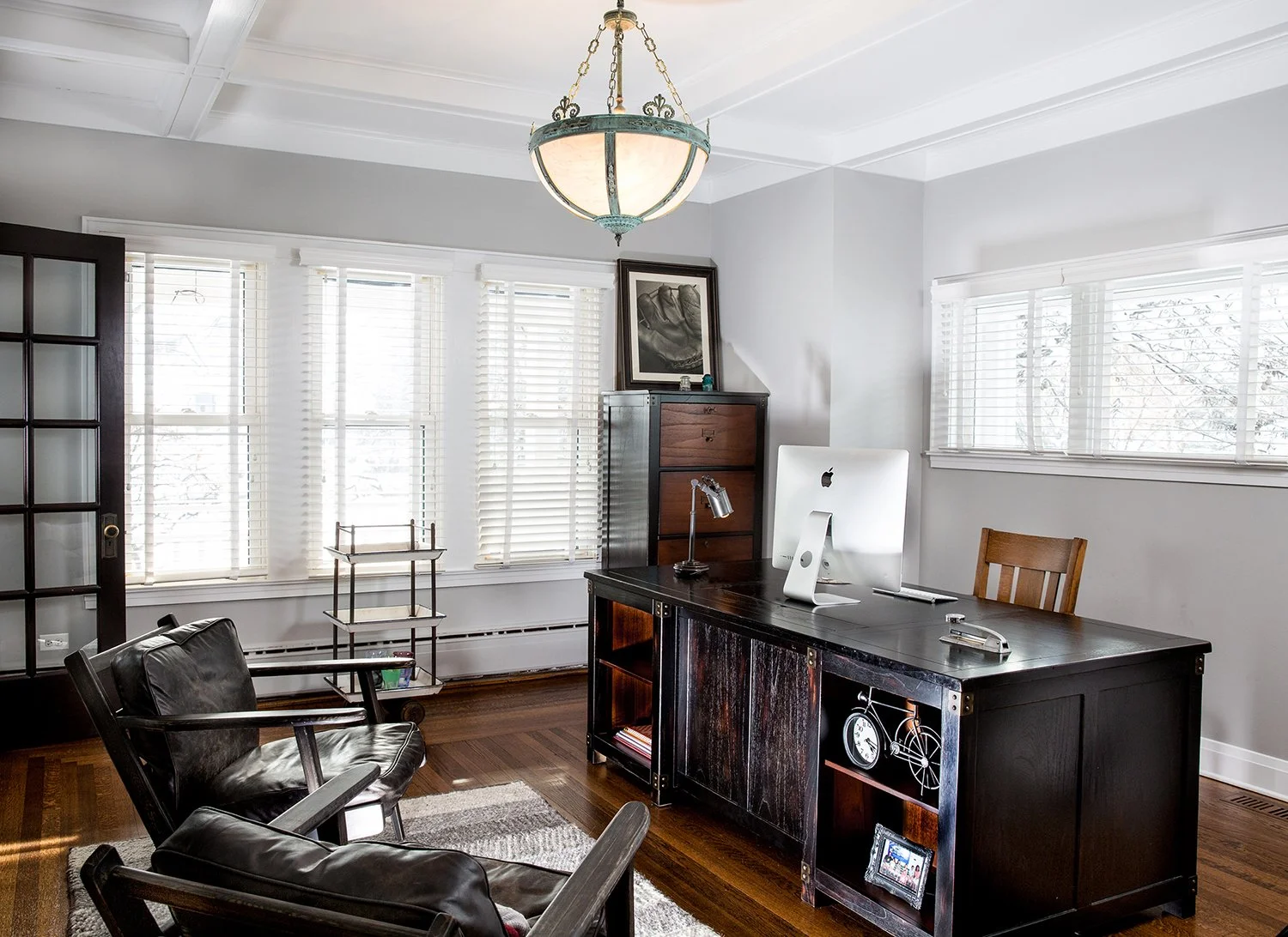 Bright Home Office with Dark Wood Desk, An Apple iMac, Two Leather Armchairs, a Coffered Ceiling, and a Vintage Pendant Light