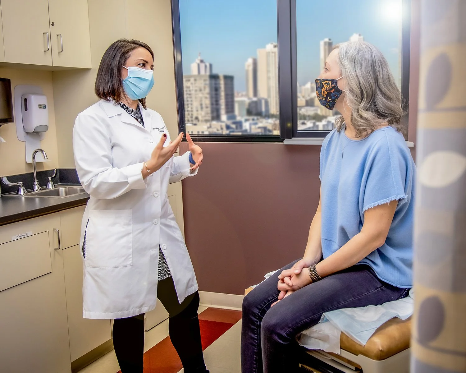 Female Doctor in White Lab Coat and Mask talking to Female Patient in her 50's in Modern Clinical Setting with Big Window