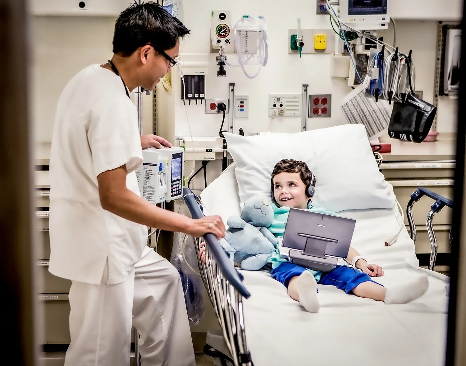 Doctor Checking on Young Patient with Laptop, Headphones and Stuffed Animal in Hospital Room.