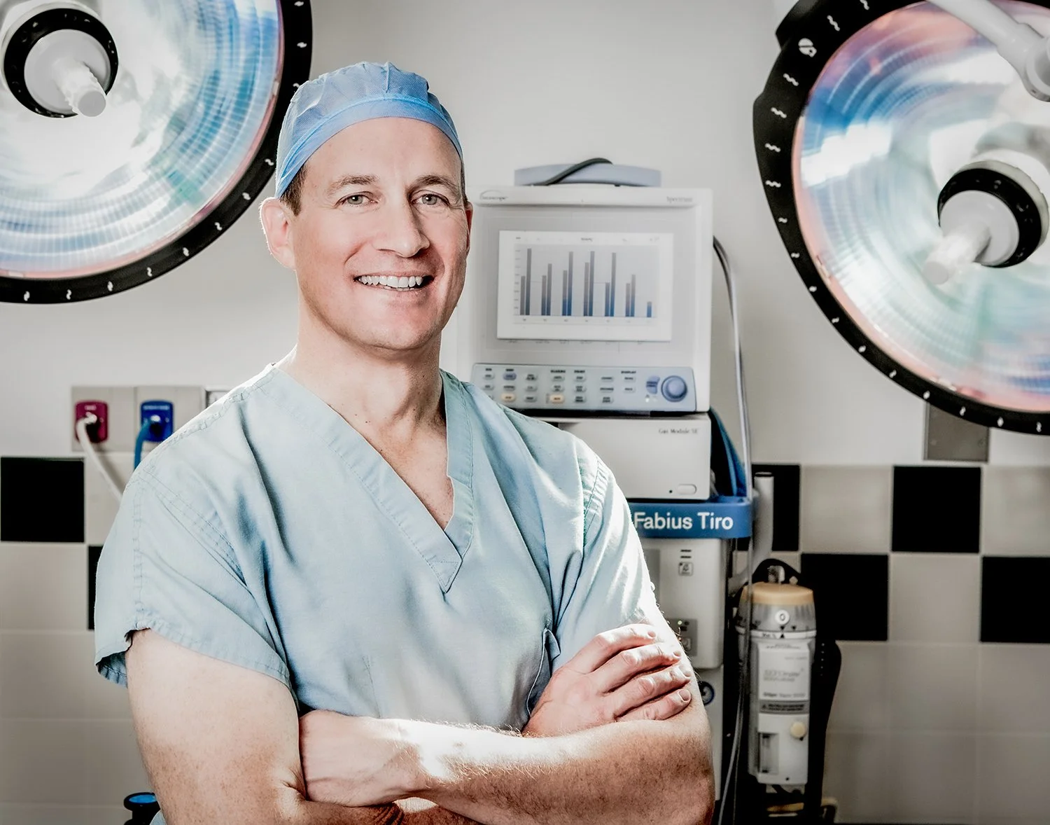 Portrait of Male Doctor in Blue Scrubs and Hair Net in Front of Large Medical Lights and Computer Screen.