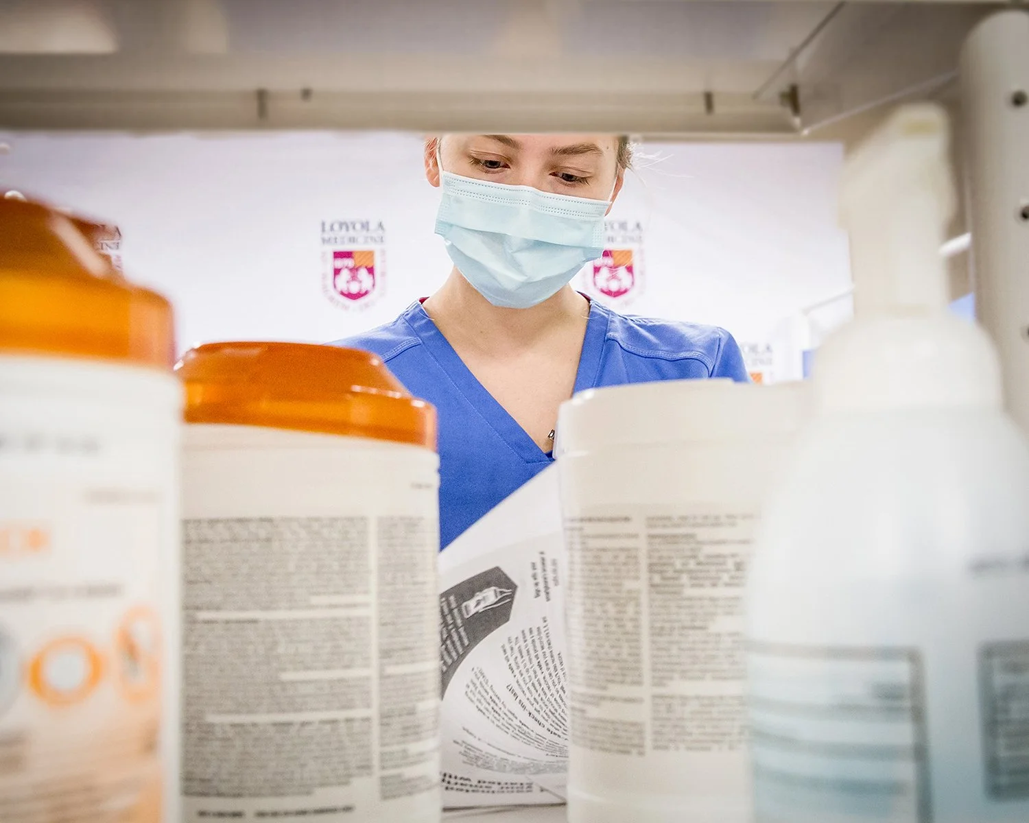 Female Nurse in Blue Scrubs and Mask Looking at Paperwork while Searching Through a Cabinet for Medical Supplies
