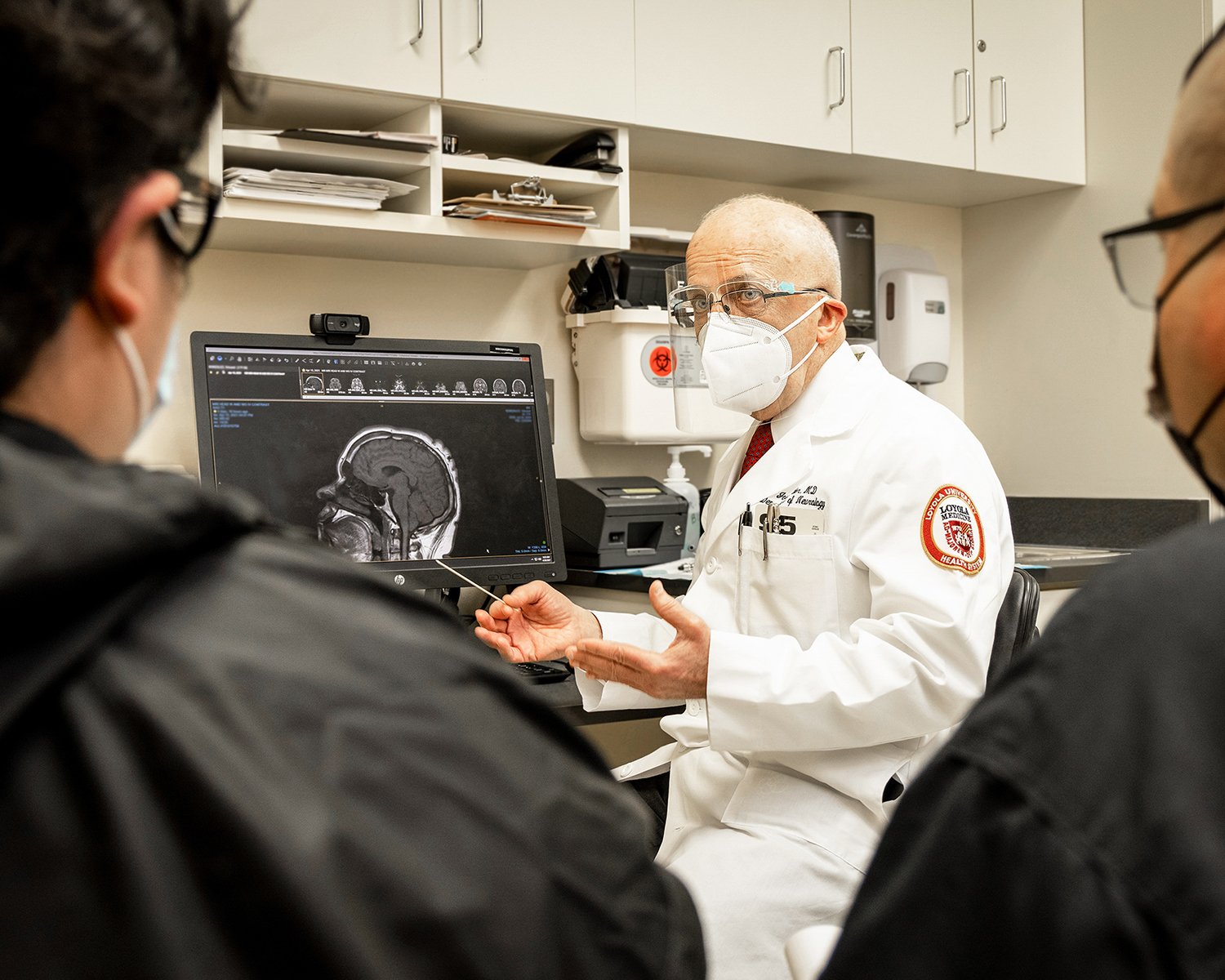 Male Doctor in White Lab Coat and Mask Explaining Brain Scans on a Computer to a Male Patient and his Father