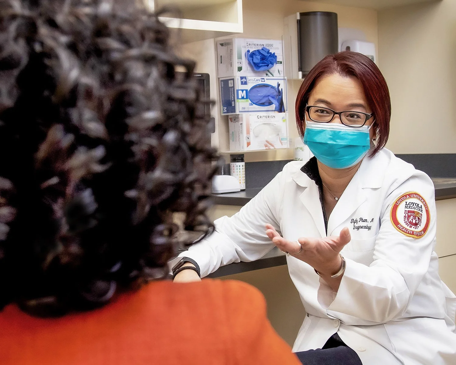 Female Nurse with White Lab Coat and Mask Talking and Gesturing with her Hand to Female Patient in Clinical Setting
