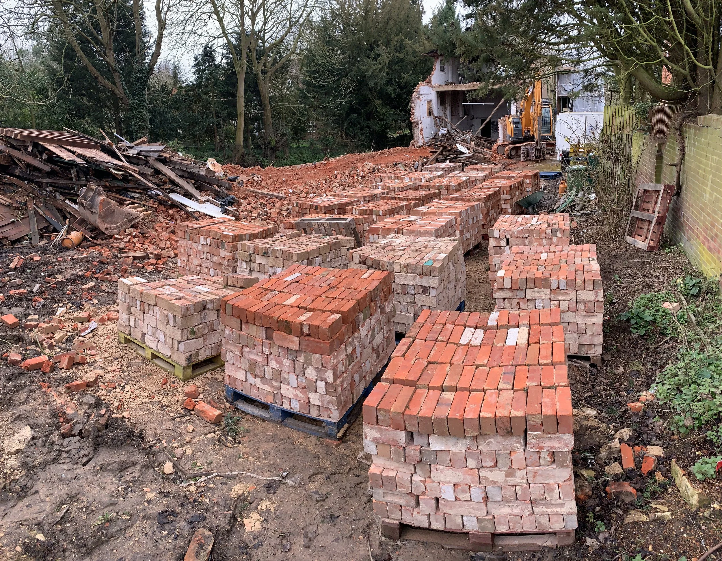 Construction site with a partially demolished building, bricks, a small excavator, and piles of debris surrounded by trees and a fence.
