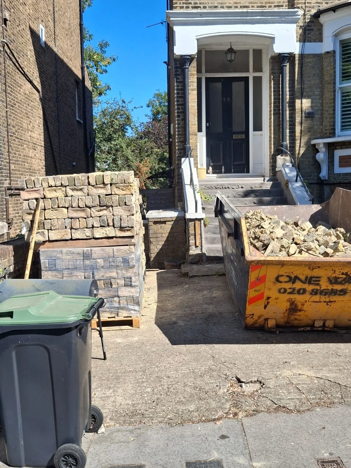Construction site with a staircase leading to a house entrance, a large dumpster filled with rocks, a pallet of bricks, and a trash bin on a city street.