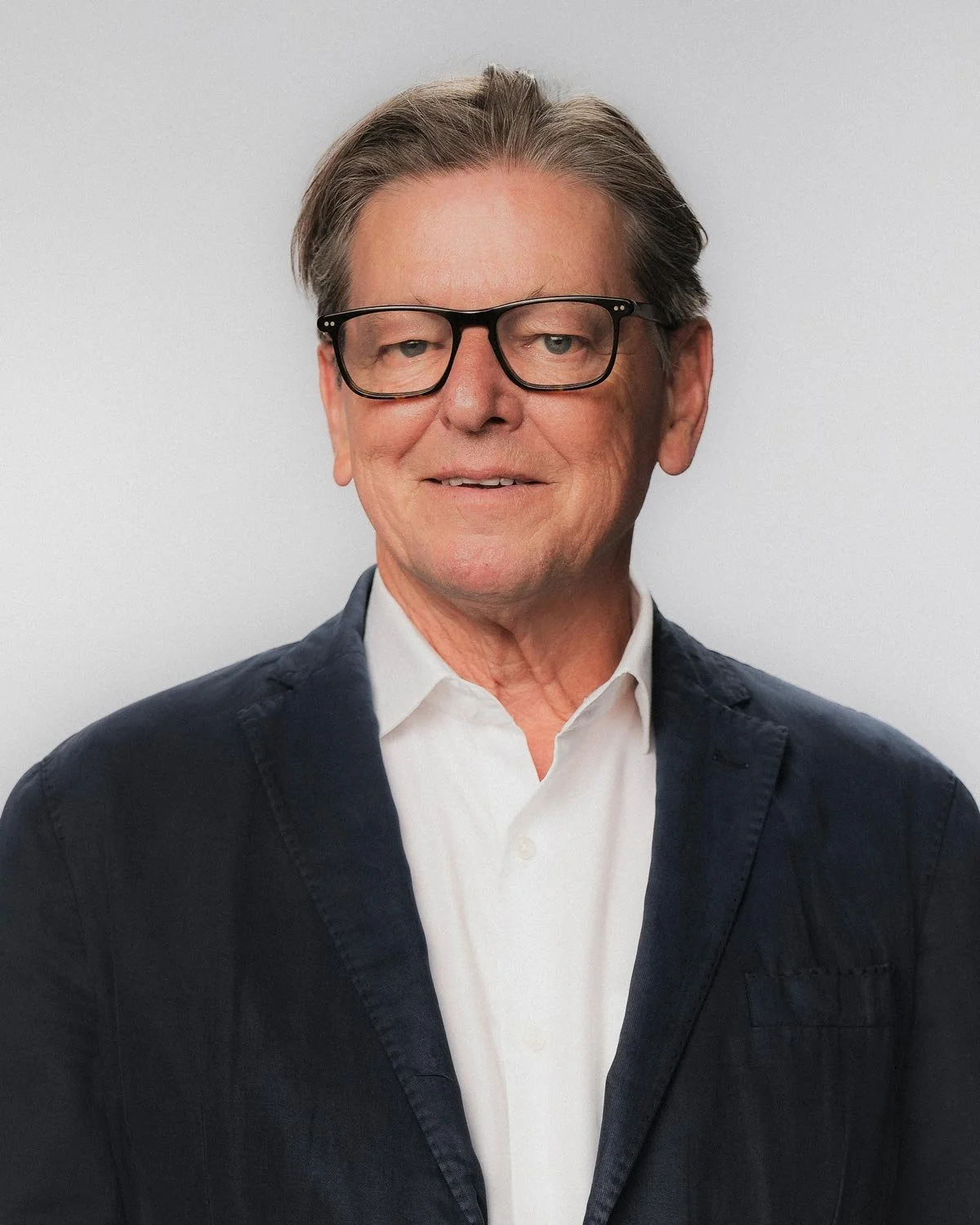 Thomas Hotko mit dunklen Haaren, Brille, in einem weißen Hemd und dunklem Sakko vor einem grauen Hintergrund.