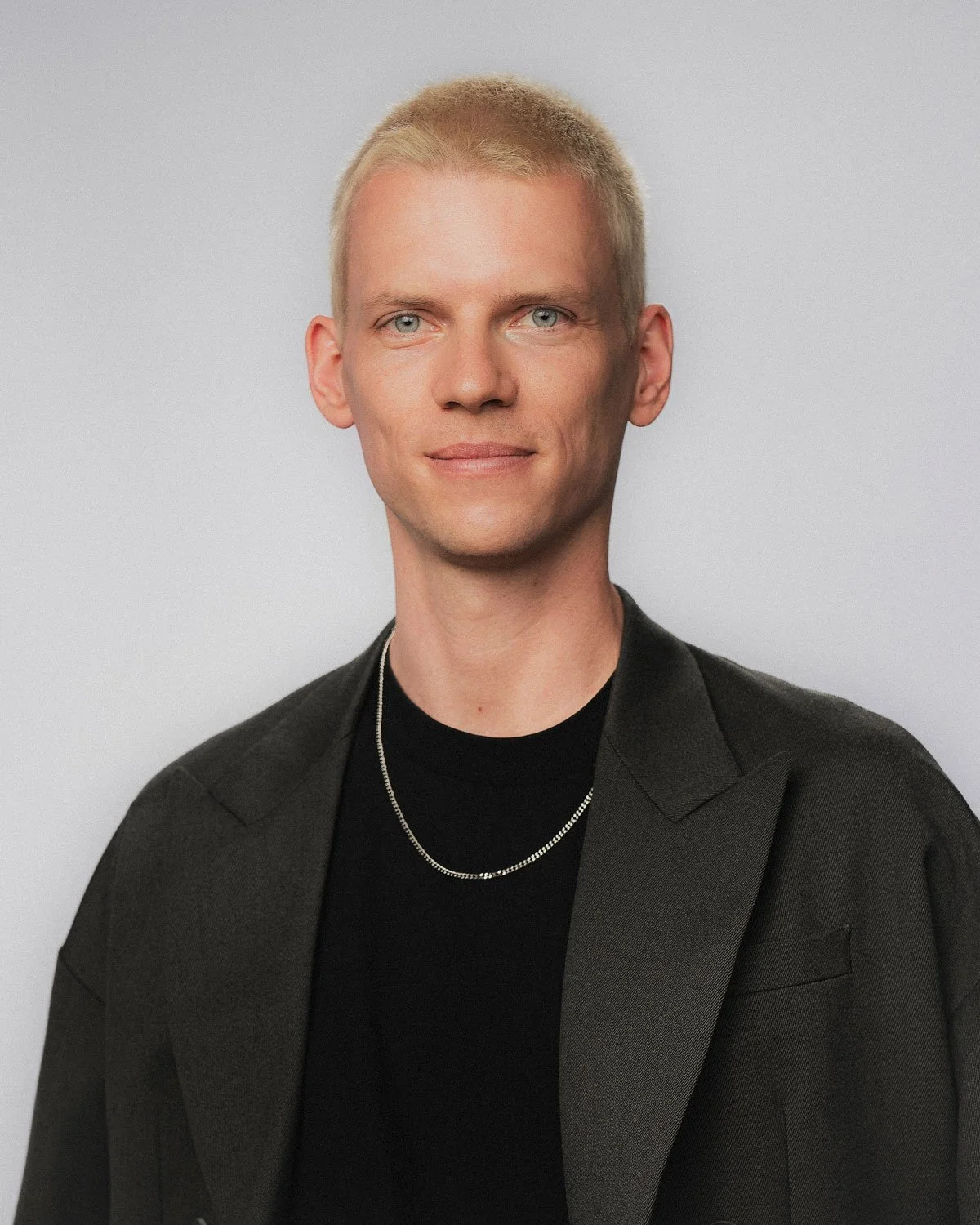 Designer Christian Leban mit blondem Kurzhaarschnitt, dunklem Anzug und schwarzem T-Shirt, trägt eine Silberkette, vor einem hellgrauen Hintergrund.