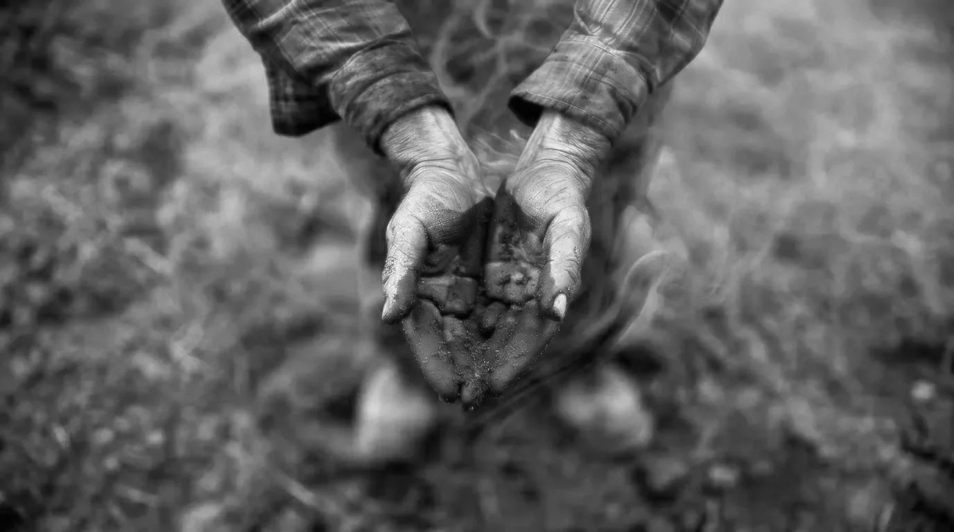 Black and white photo of a person holding wet dirt or soil in their hands, with the soil forming a small mound, in an outdoor setting.