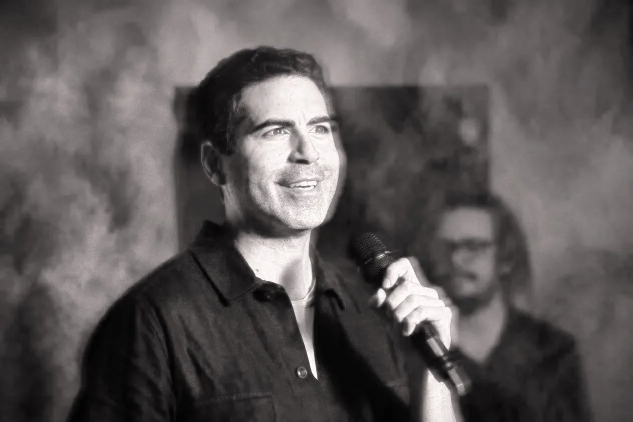 A man holding a microphone and smiling, with a woman with glasses blurred in the background.