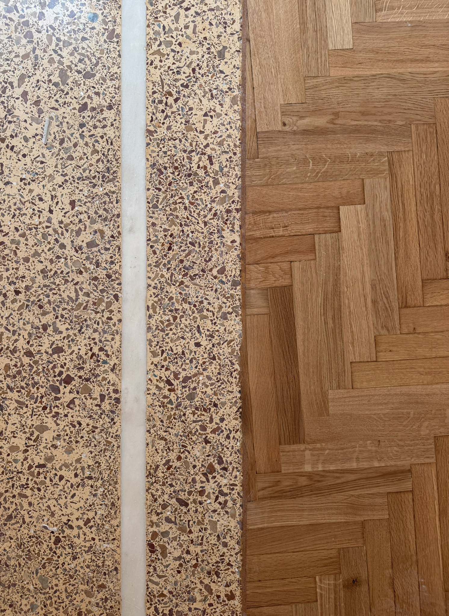 Close-up image showing a transition between terrazzo floor and wood parquet floor with a white strip in between.