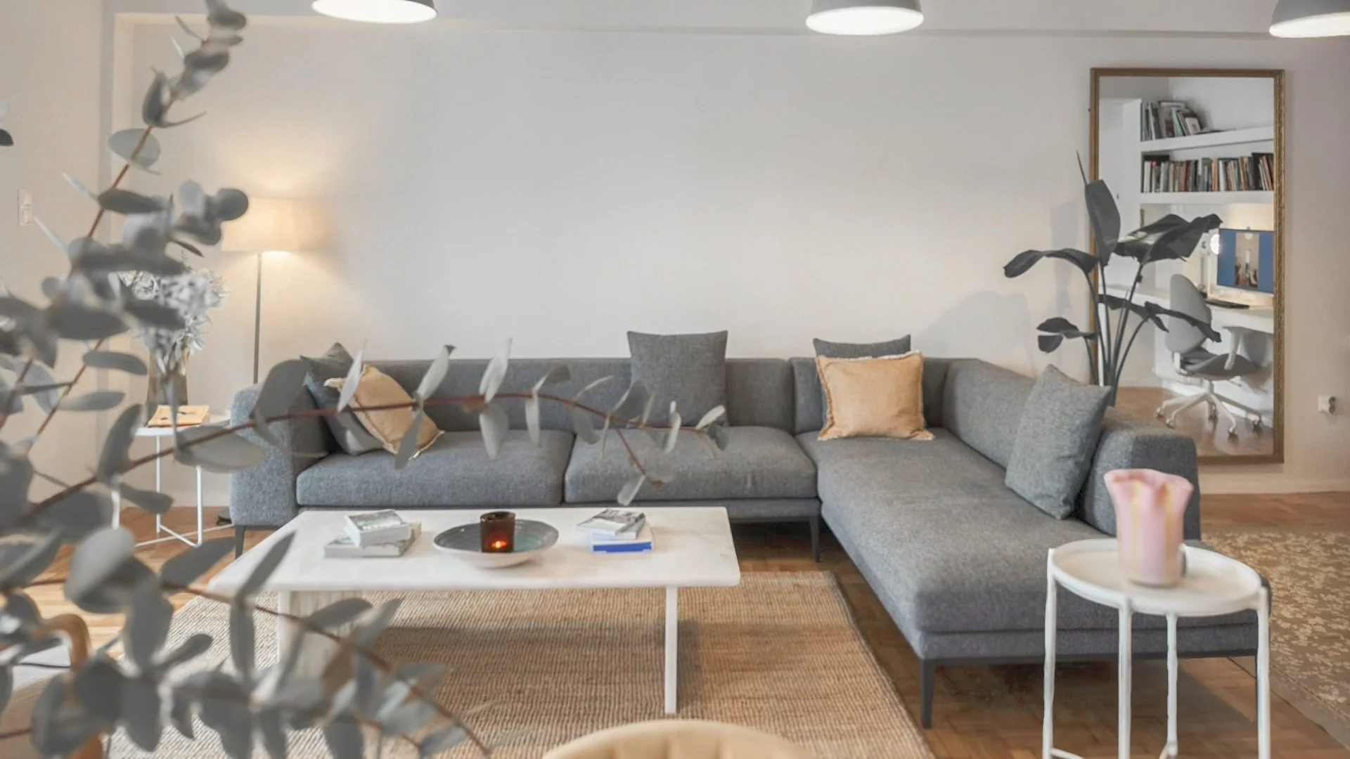Living room with a gray sectional sofa, beige and gray pillows, coffee table with books and a candle, large mirror on the wall, indoor plants, and a visible workspace with a computer and books in the background.