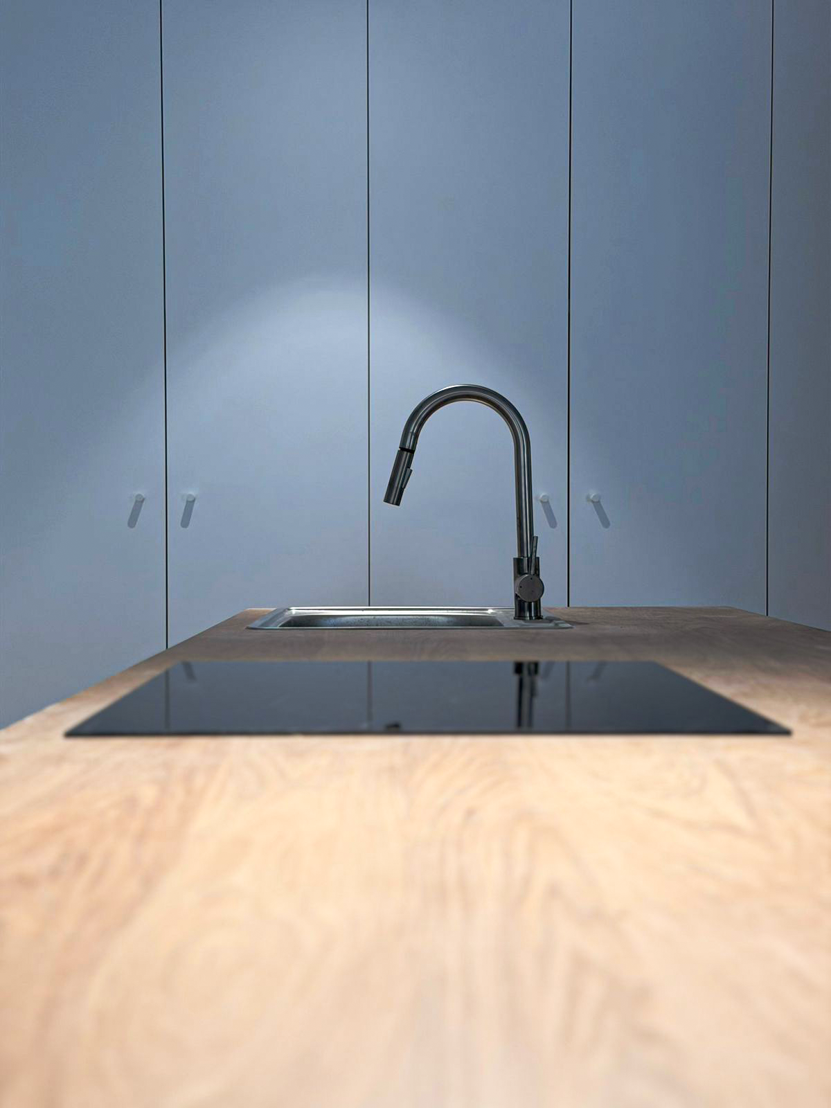 Modern kitchen with a black stovetop and a stainless steel sink with a tall, curved faucet, set against a pale blue wall with cabinets.