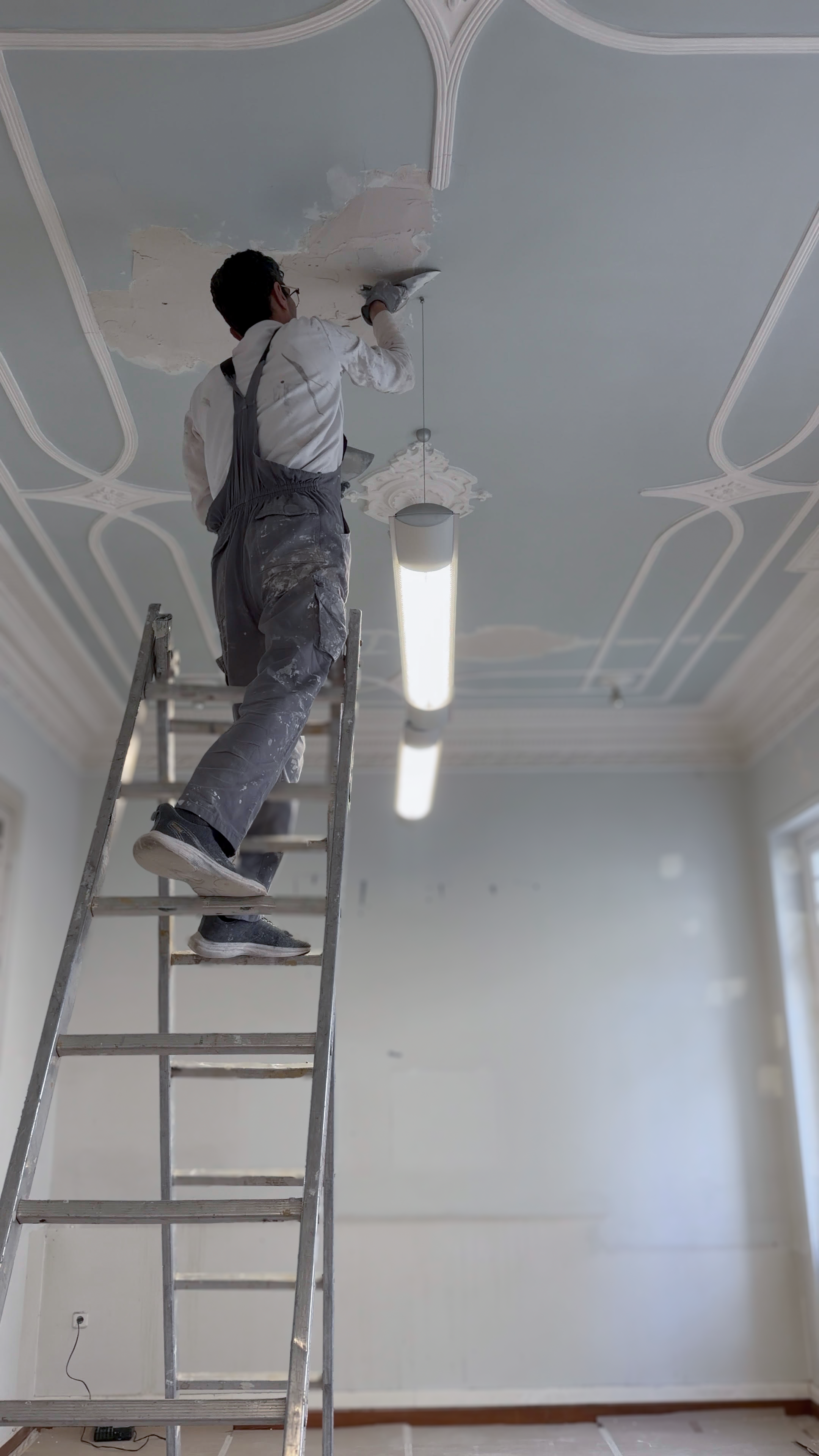 A person standing on a ladder repairing a damaged ceiling with paint and tools. The ceiling has decorative molding and a hanging light fixture.
