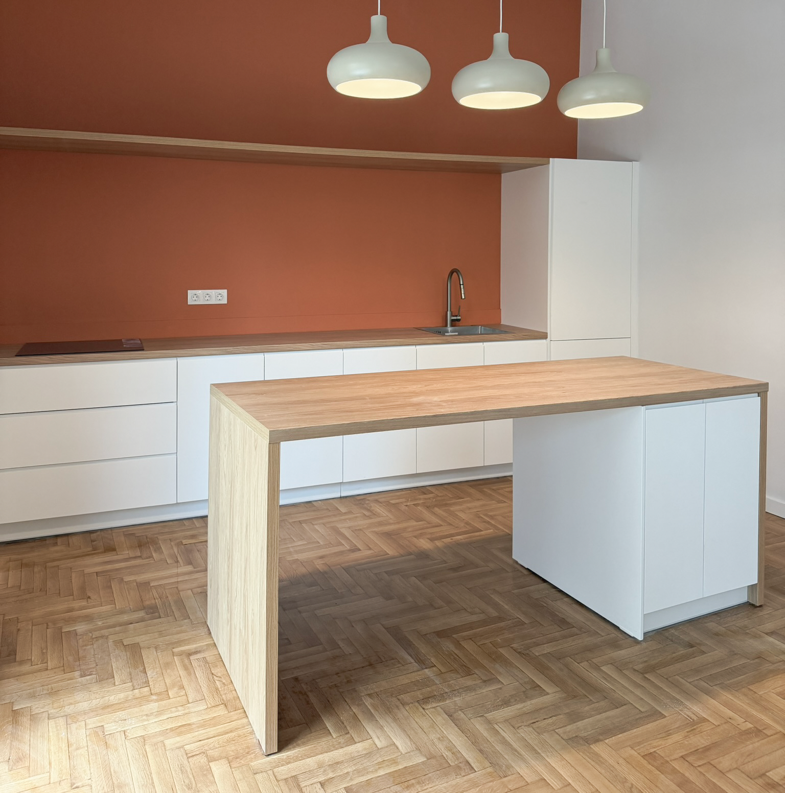 Empty modern kitchen with white cabinets, a wooden countertop, a sink with a silver faucet, and three white pendant lights hanging from the ceiling. The wall is painted terracotta, and the floor is wooden parquet.