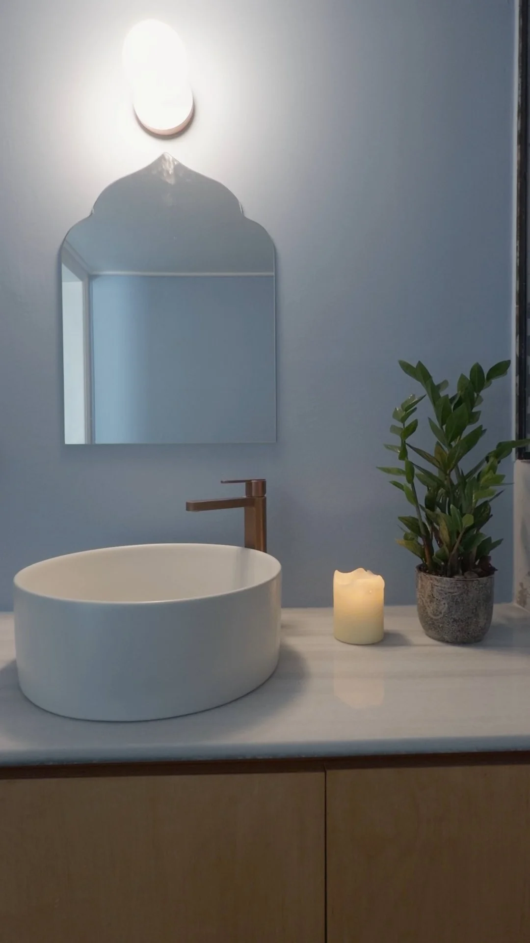 Bathroom countertop with a white vessel sink, a rose gold faucet, a lit candle, a green potted plant, a mirror, and a wall-mounted light