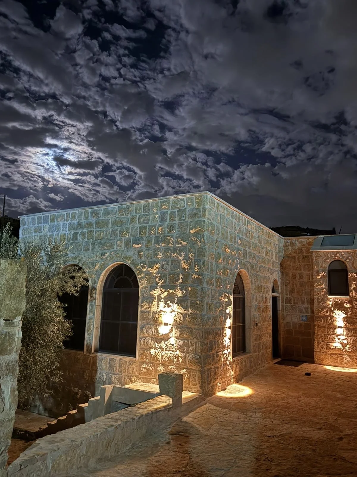 Stone house illuminated by exterior lights under a night sky with clouds and the moon.