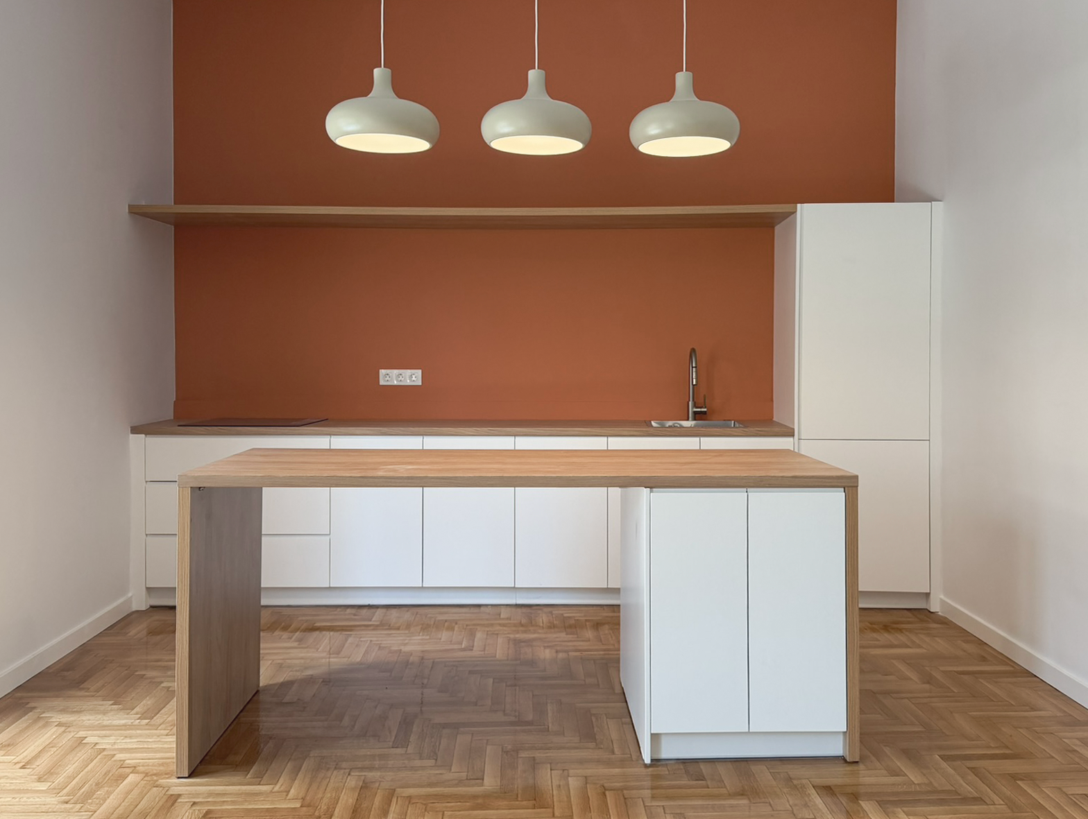 Minimalist kitchen with white cabinets, a small wooden kitchen island, a rust-colored accent wall, three hanging white pendant lights, and a wooden sink counter.