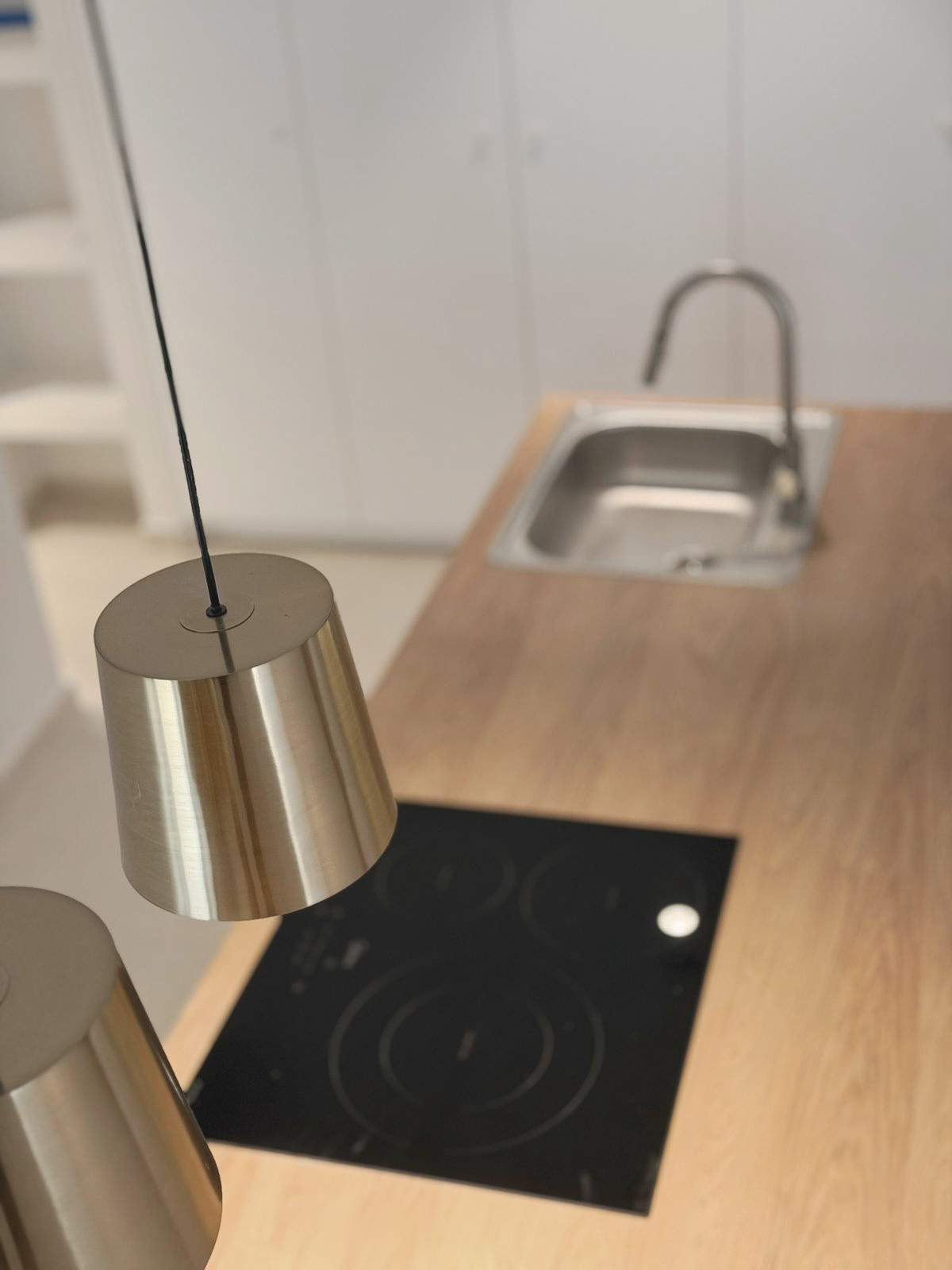 Close-up of a modern kitchen with a black electric stove on a wooden countertop, a stainless-steel sink with a faucet, white cabinets in the background, and a metallic pendant light fixture hanging above.