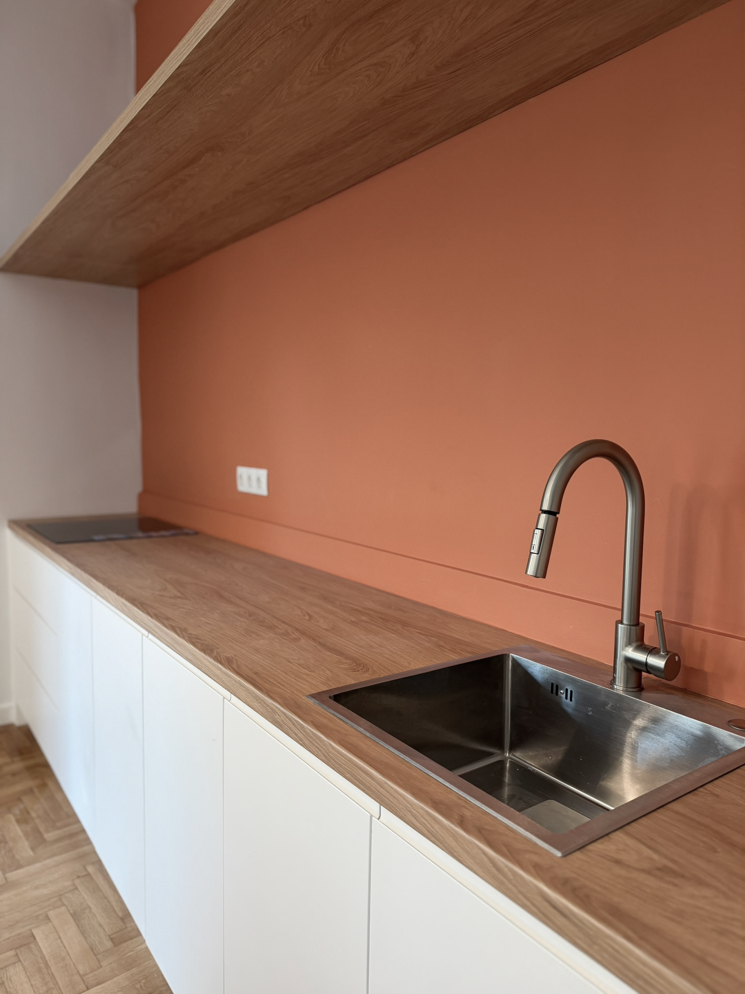 Modern kitchen with a wooden countertop, a stainless steel sink, and a sleek faucet against a terracotta-colored wall with electrical outlets.