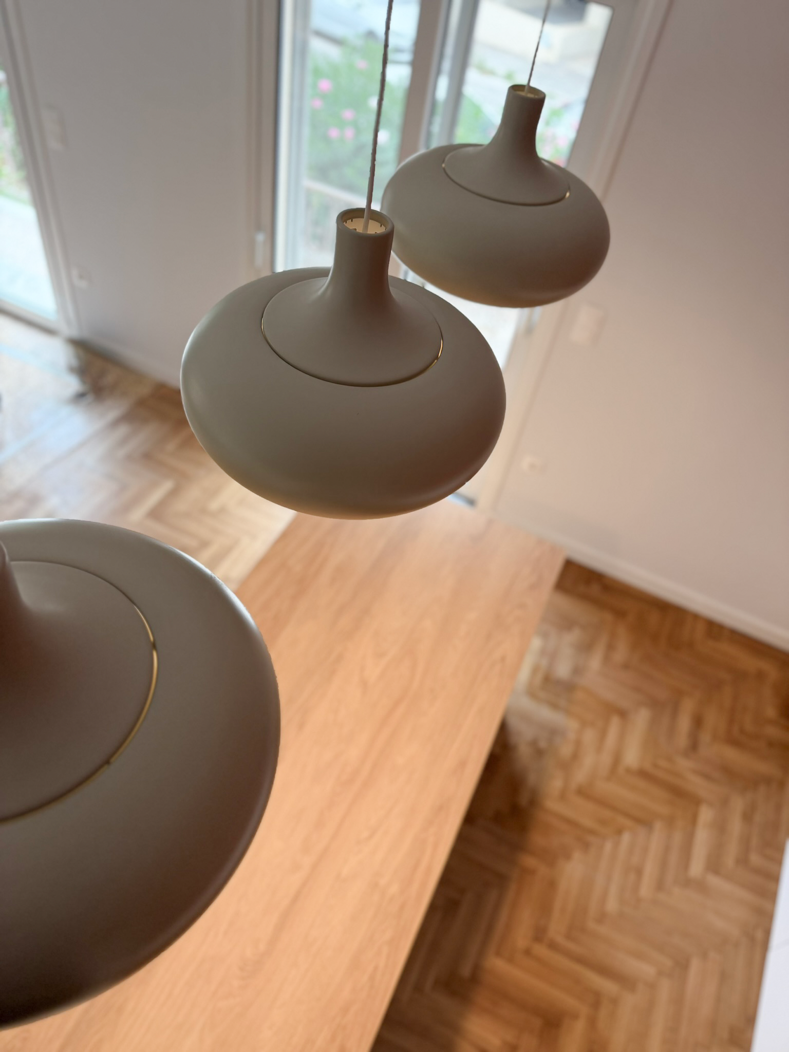 Two modern white pendant light fixtures hanging in a room with a wooden floor and a glass door showing a garden outside.