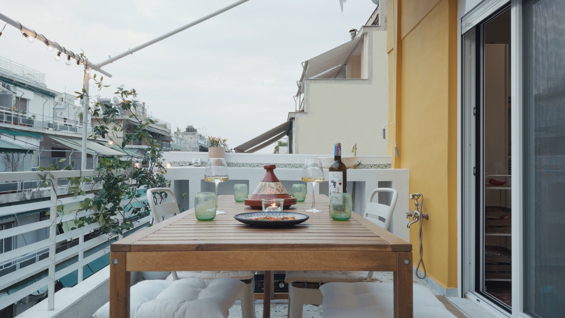Balcony dining area with wooden table set for dinner, wine glasses, green candles, wine bottle, and a pizza plate, overlooking neighboring buildings and string lights.