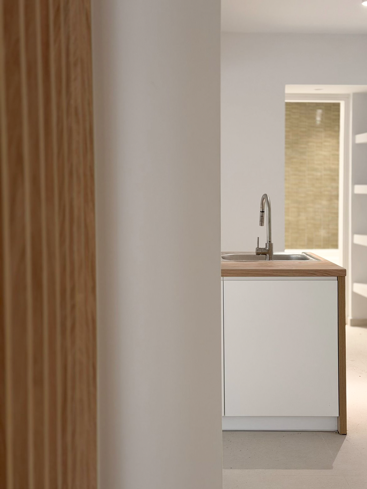 Partial view of a modern kitchen with a white cabinet, wooden countertop, and a stainless steel faucet, seen through a doorway with a wooden frame.