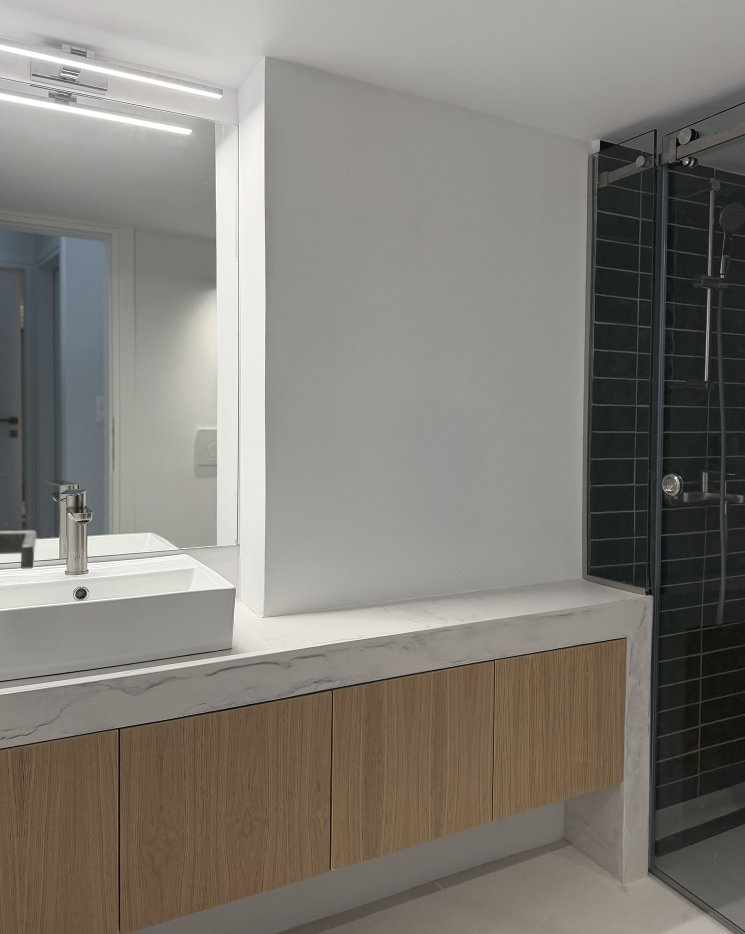 Modern bathroom vanity with a rectangular white sink and a sleek metallic faucet, wooden cabinet below, and a large mirror above. Part of a glass shower enclosure with dark tiles is visible on the right.