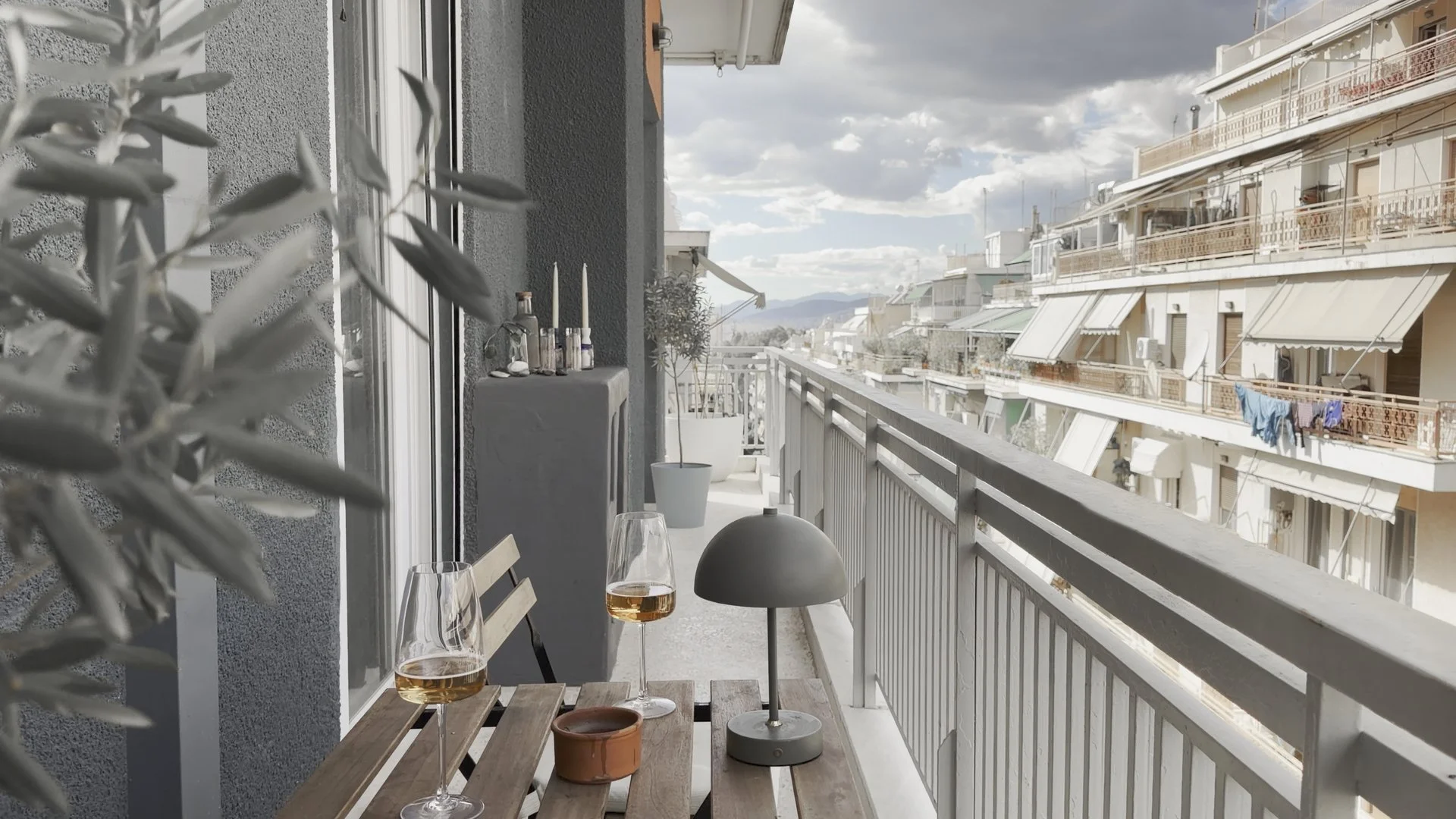 Balcony with two glasses of wine on a wooden table, potted plants, and a modern lamp, overlooking neighboring apartment buildings with laundry hanging on the balconies under a partly cloudy sky.