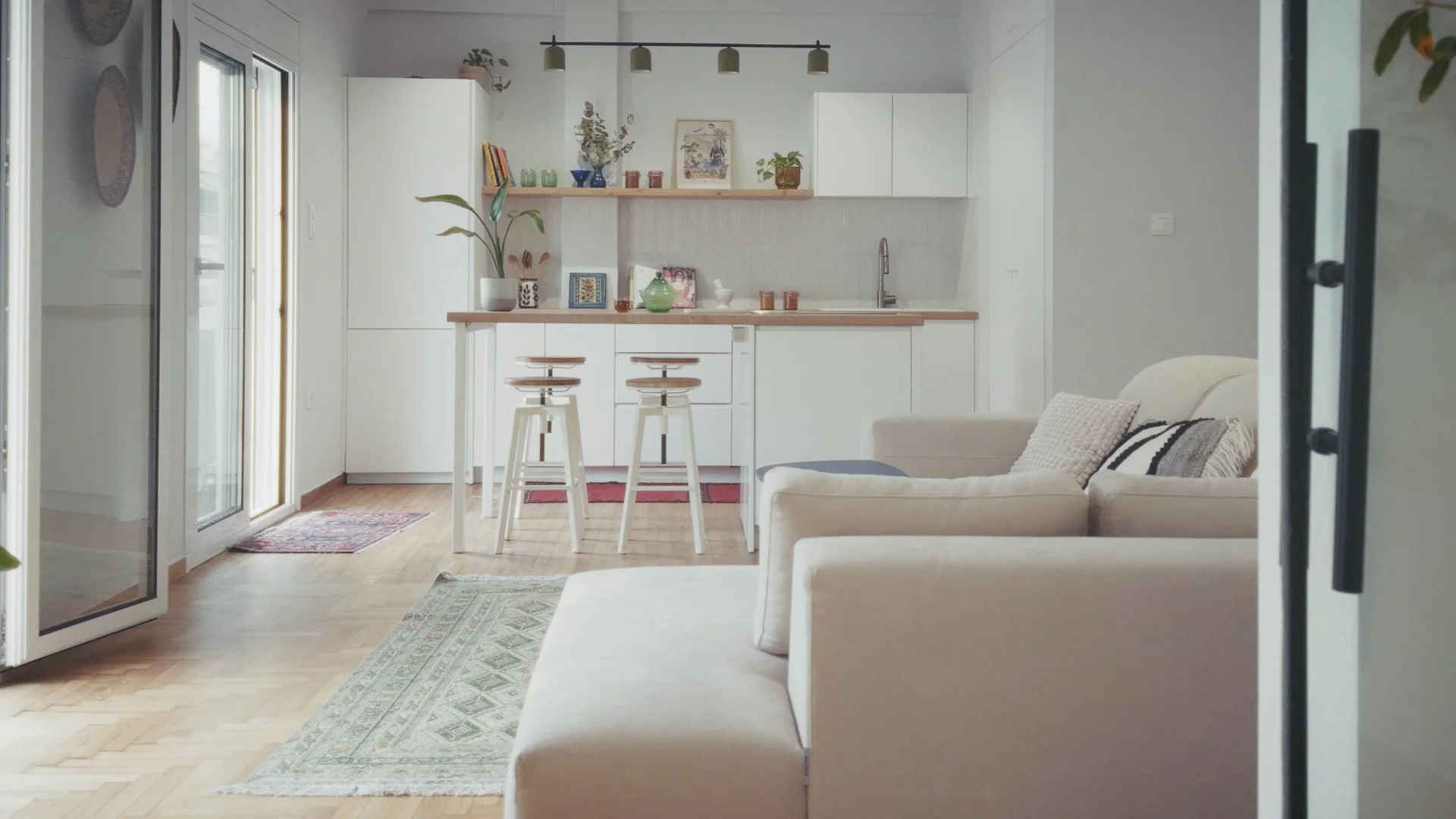 Open-concept living room and kitchen with white cabinetry, a small dining area, and a beige sofa, illuminated by natural light from sliding glass doors.