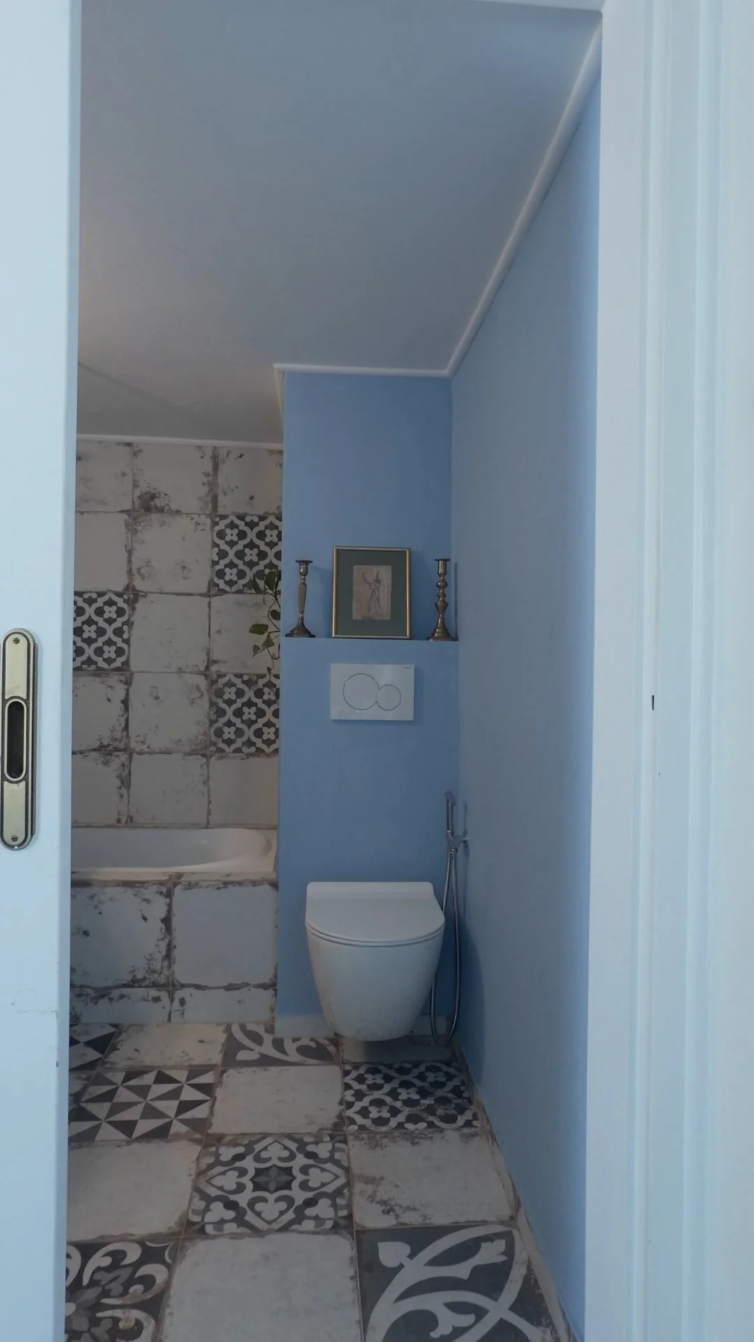 Small bathroom with a modern toilet, blue painted walls, and a decorative tiled floor. A small niche on the wall contains framed pictures and candlesticks.