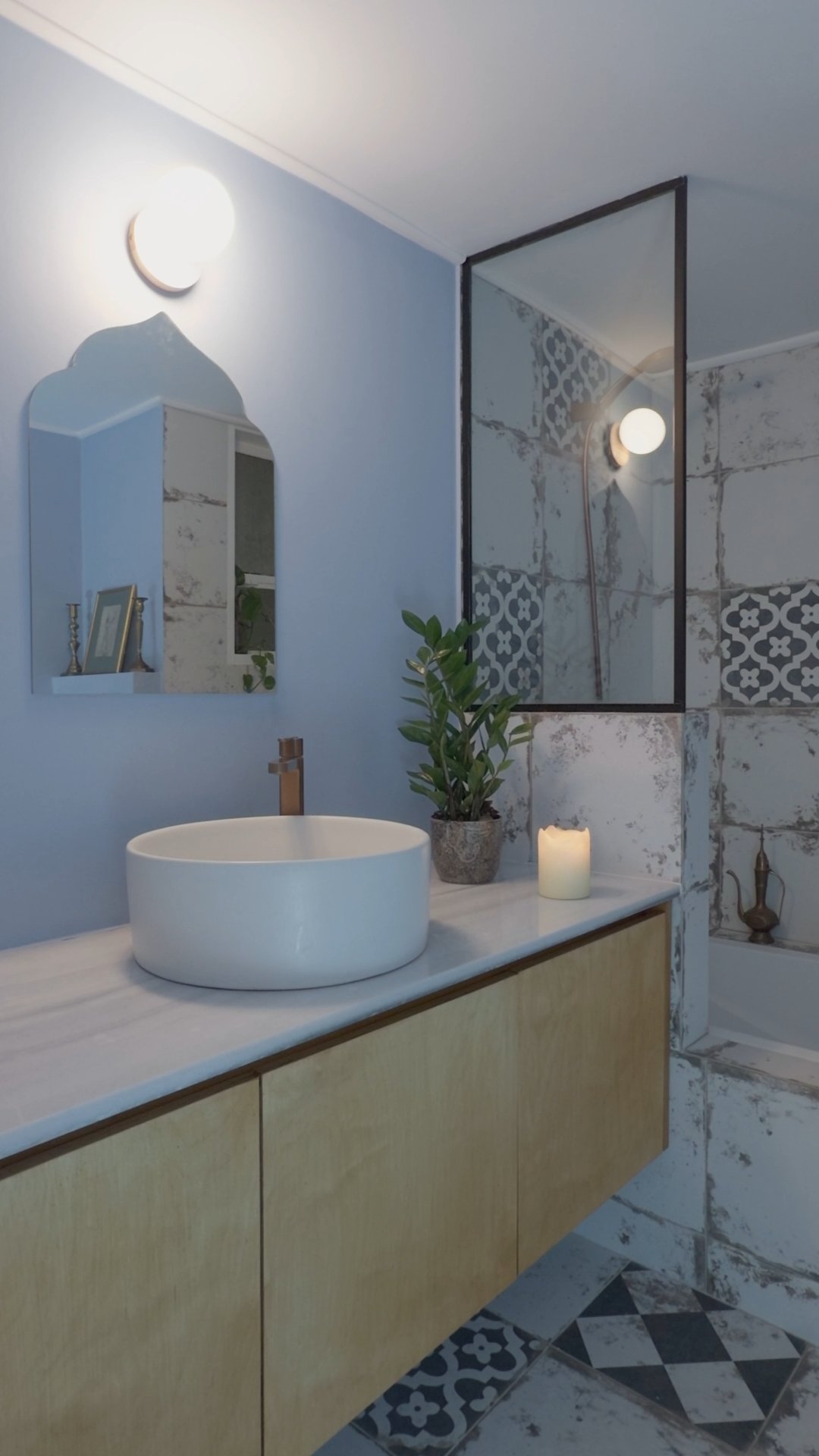 Modern bathroom vanity with a round white vessel sink, a copper faucet, a potted plant, a lit candle, a mirror, and a decorated wall with patterned tiles and a glass shower door.