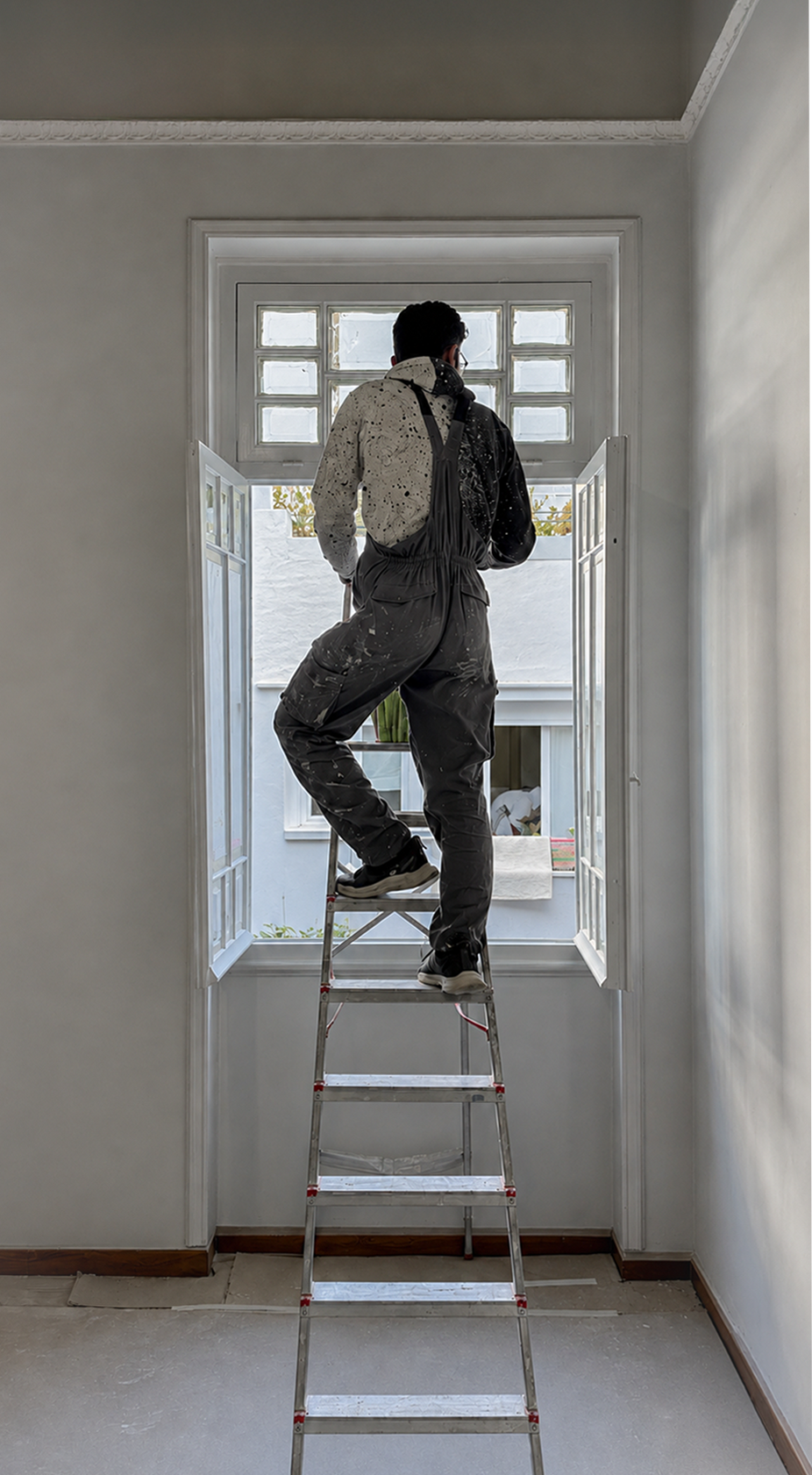 A person standing on a ladder looking out of an open window. The person is dressed in gray coveralls with paint splatters and appears to be painting or renovating the window frame.