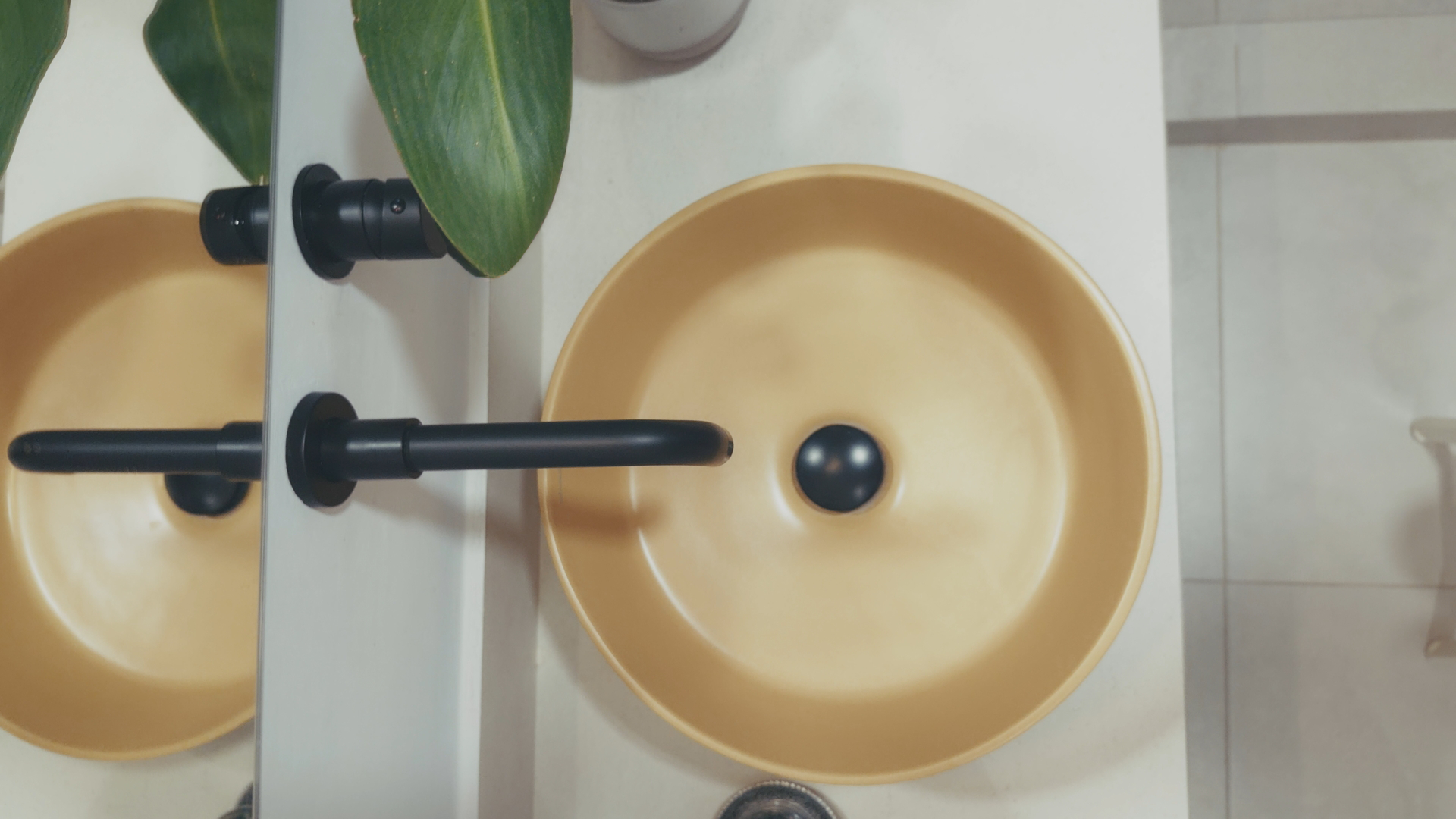 A beige bathroom sink with a black spherical drain stopper, black faucet, wall mirror, and a green plant with large leaves on the countertop.