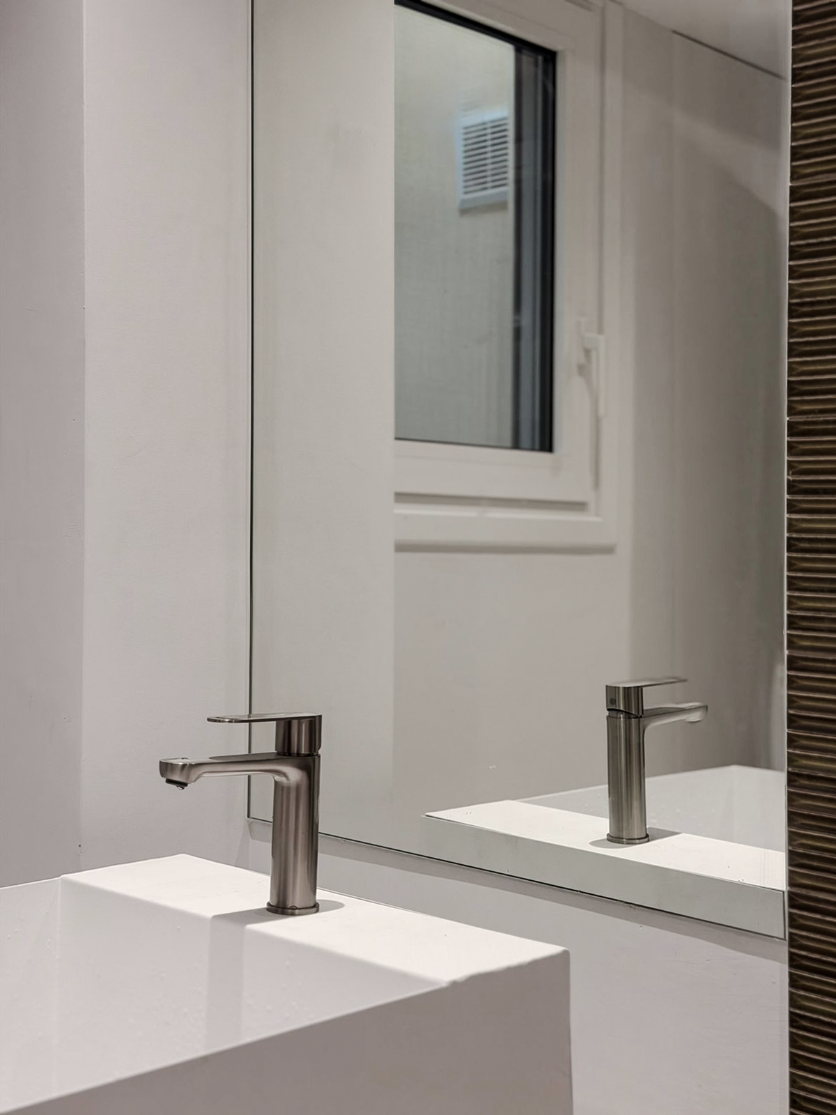 Bathroom sink with a silver faucet, white countertop, large mirror, and a window reflected in the mirror.