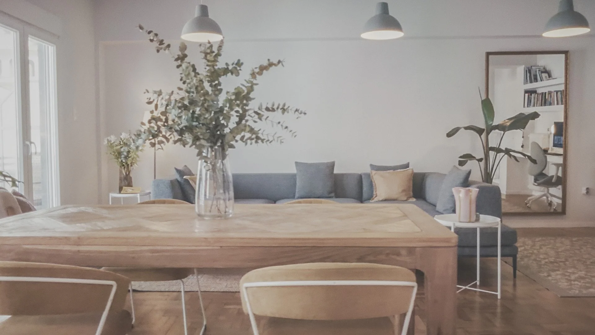 Living room with wooden dining table, decorative vase with greenery, and gray sofa with cushions. There is a large mirror on the wall reflecting the room, a bookshelf, and houseplants. Pendant lights hang from the ceiling, and sliding glass doors are