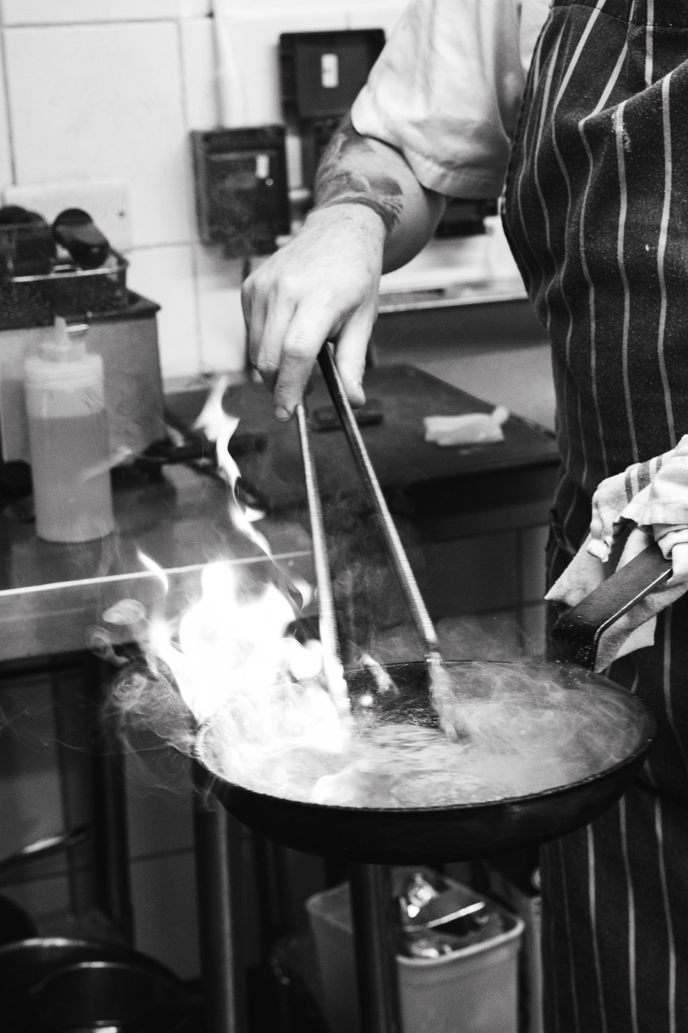 A person, wearing a striped apron, is tending to flames on a cooking surface in a kitchen. The setting includes kitchen equipment and a tiled wall.