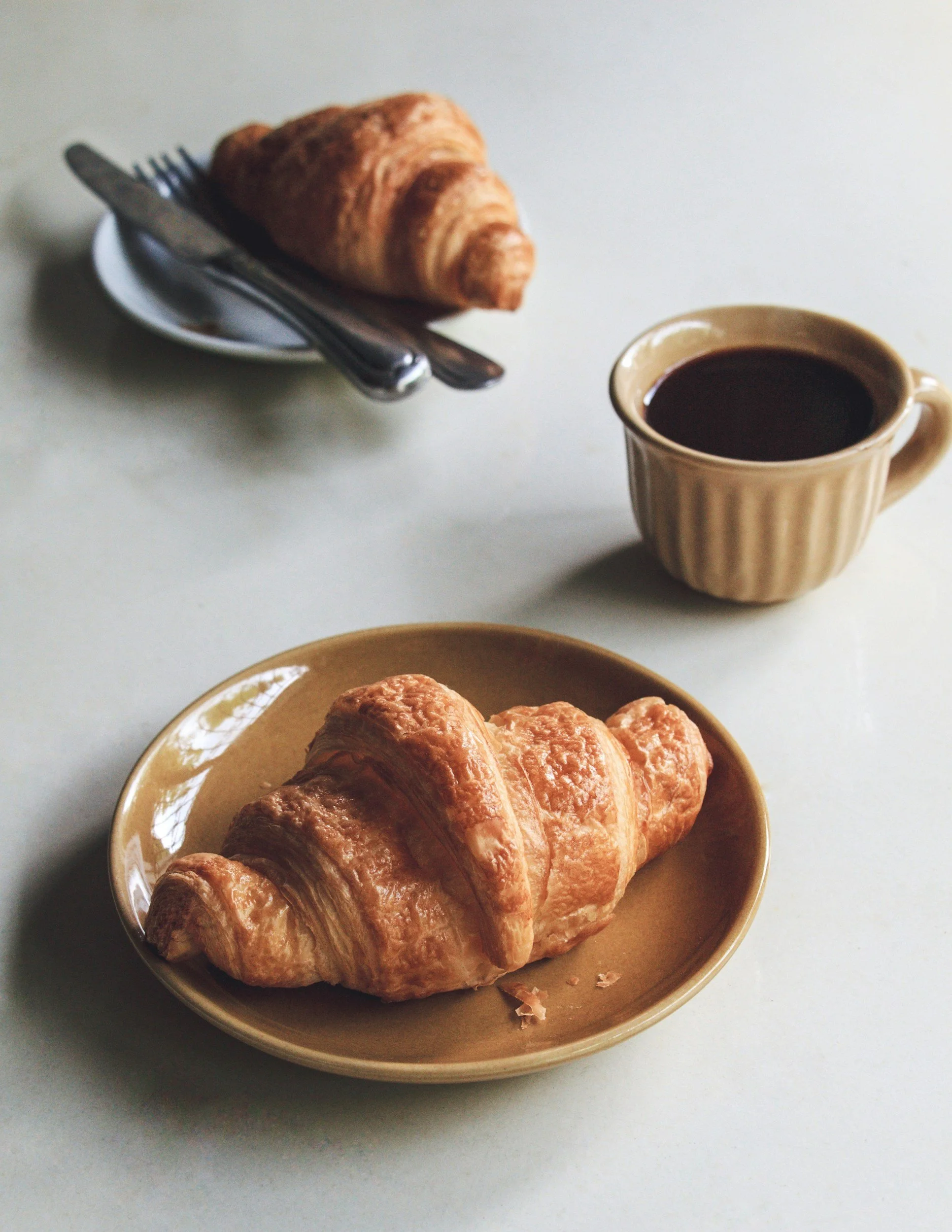 A golden brown croissant on a beige plate placed on a white surface, with a cup of black coffee next to it.