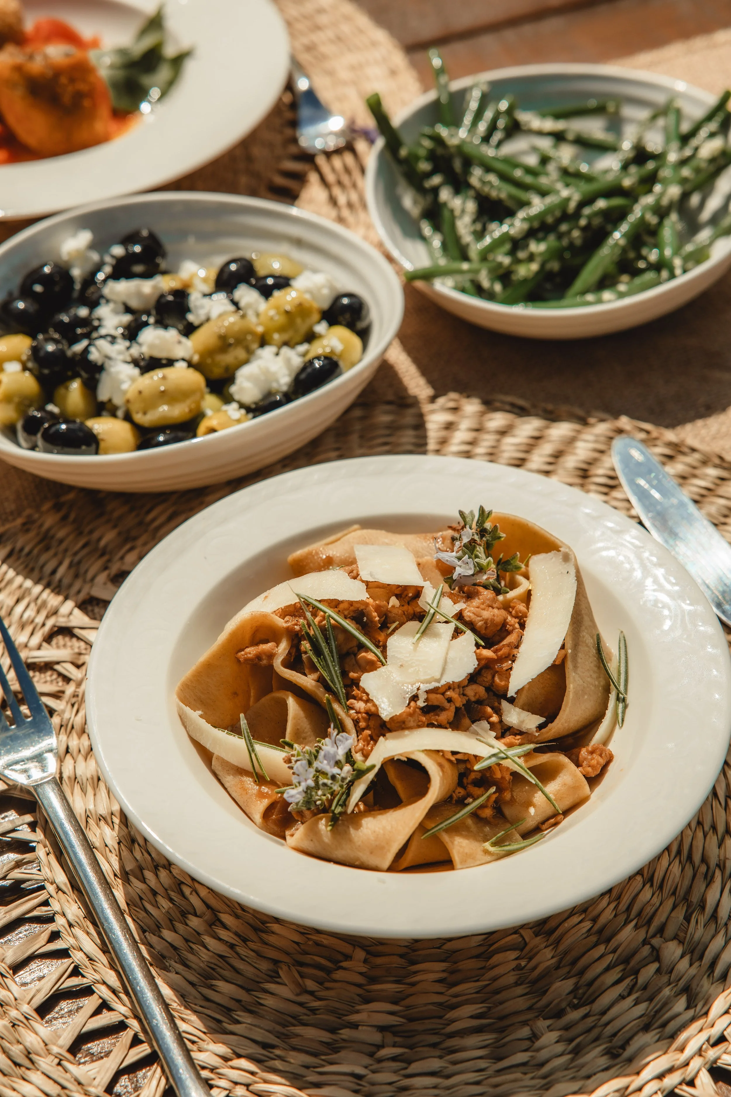 A plate of pasta with ground meat, garnished with rosemary, served on a white dish, on a woven placemat, beside a fork and knife, with additional dishes of green beans and a salad in the background.