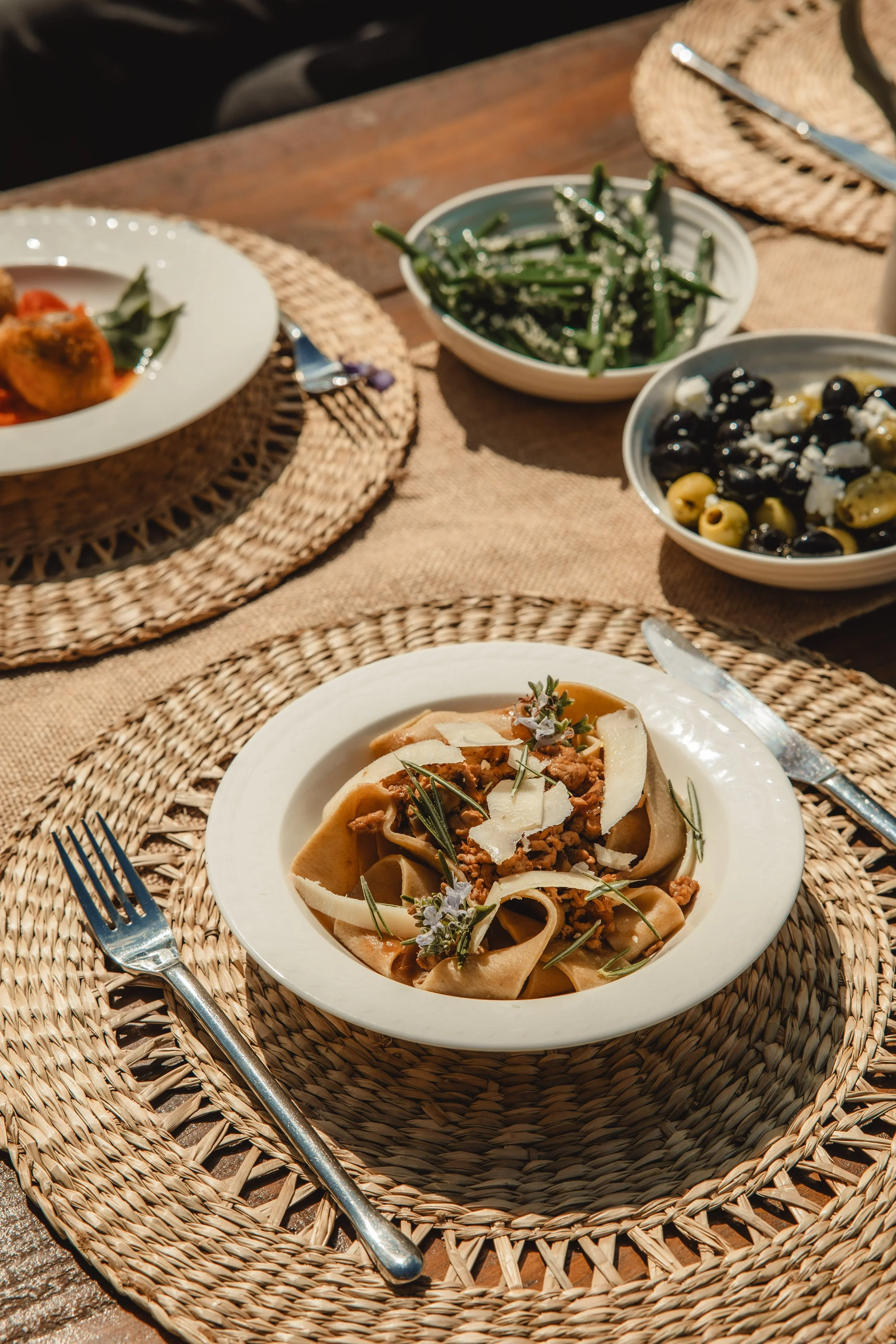A meal of pasta topped with sauce, herbs, and cheese, with a side of green beans and a bowl of a black and green olive salad on a woven placemat on a wooden table.