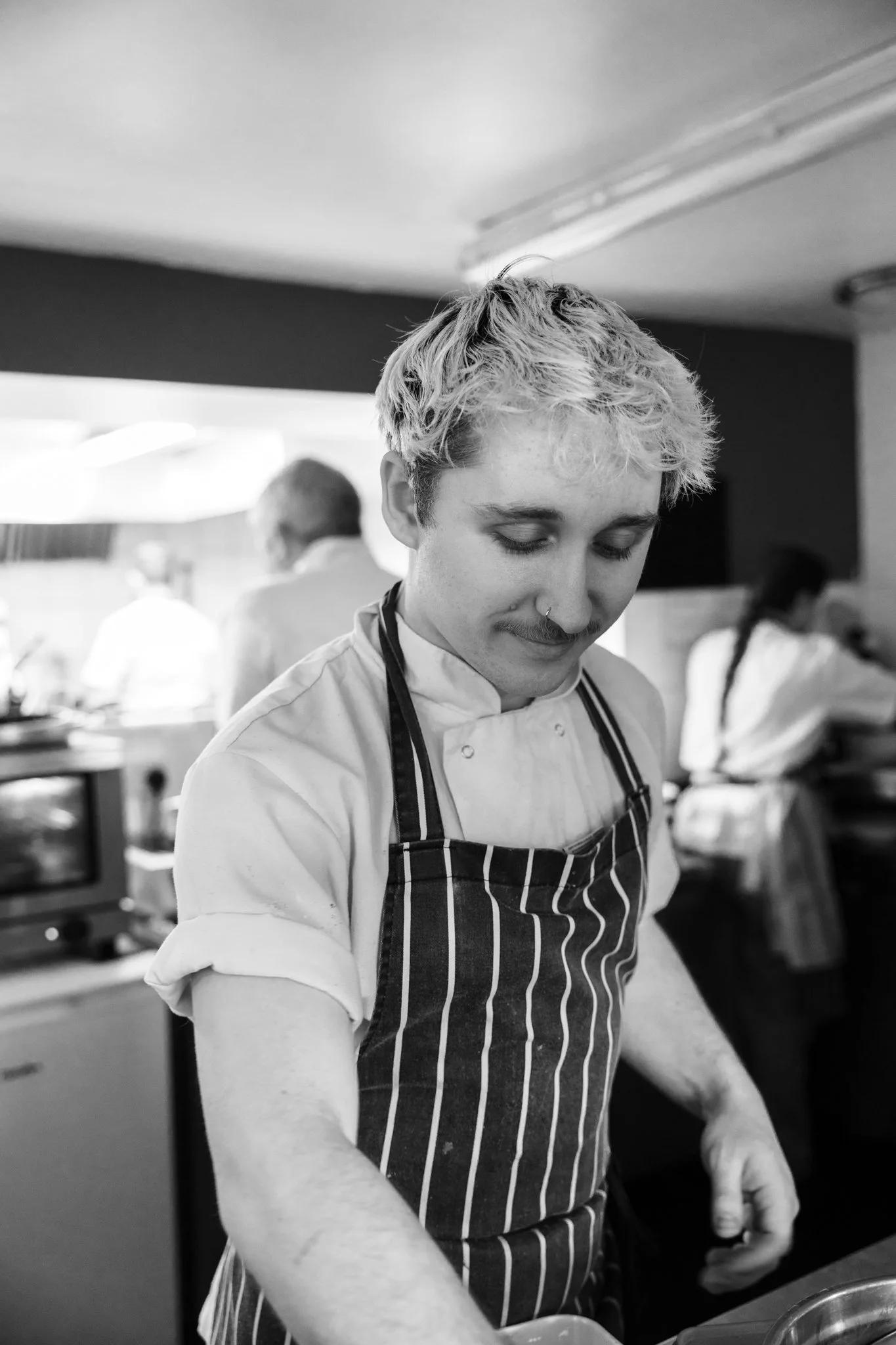 A young man with blonde, curly hair and a nose ring, wearing a striped apron and chef's coat, preparing food in a kitchen with other chefs in the background.