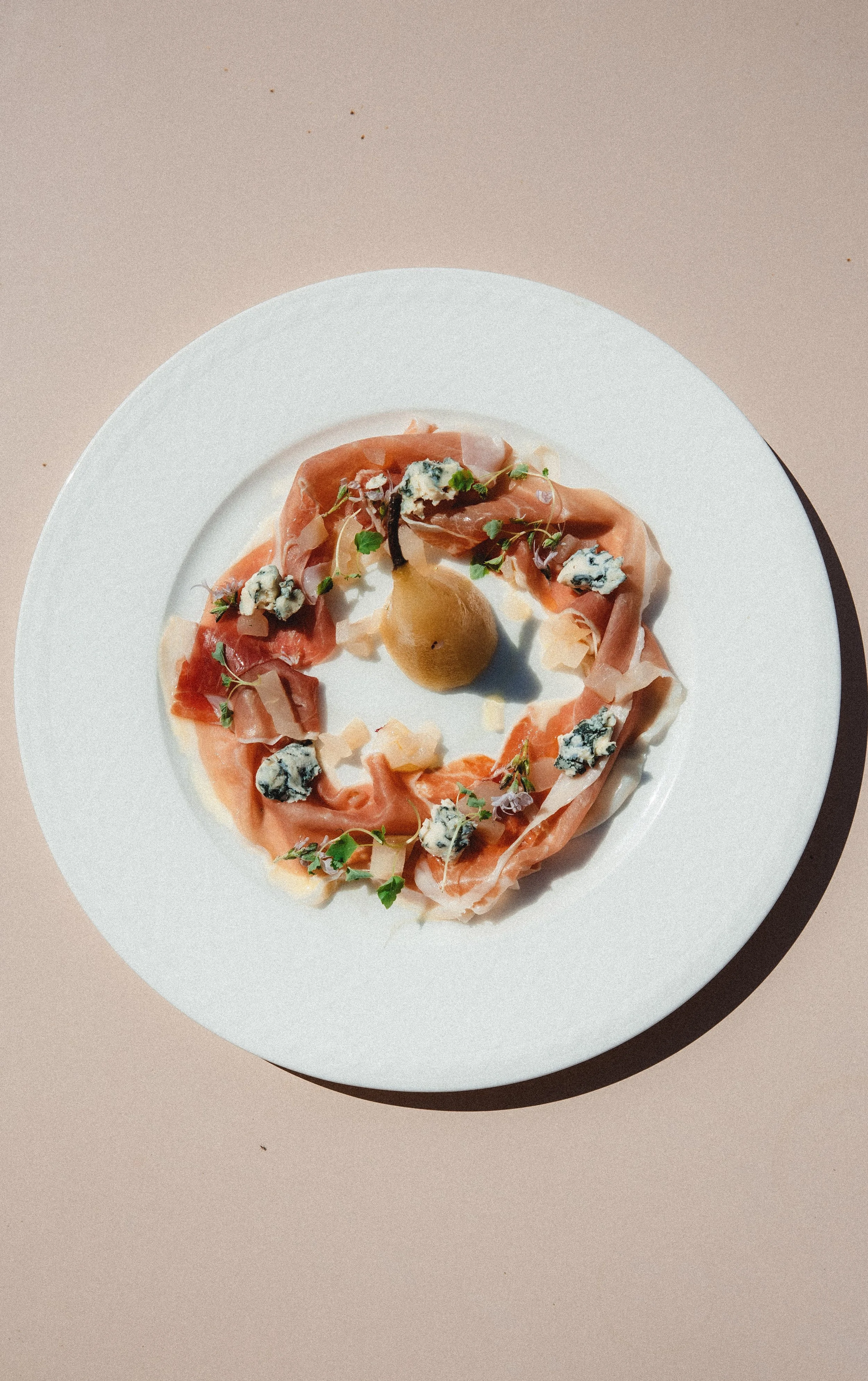 A white plate with a circular arrangement of thinly sliced prosciutto, blue cheese crumbles, fresh herbs, and a whole pear in the center.