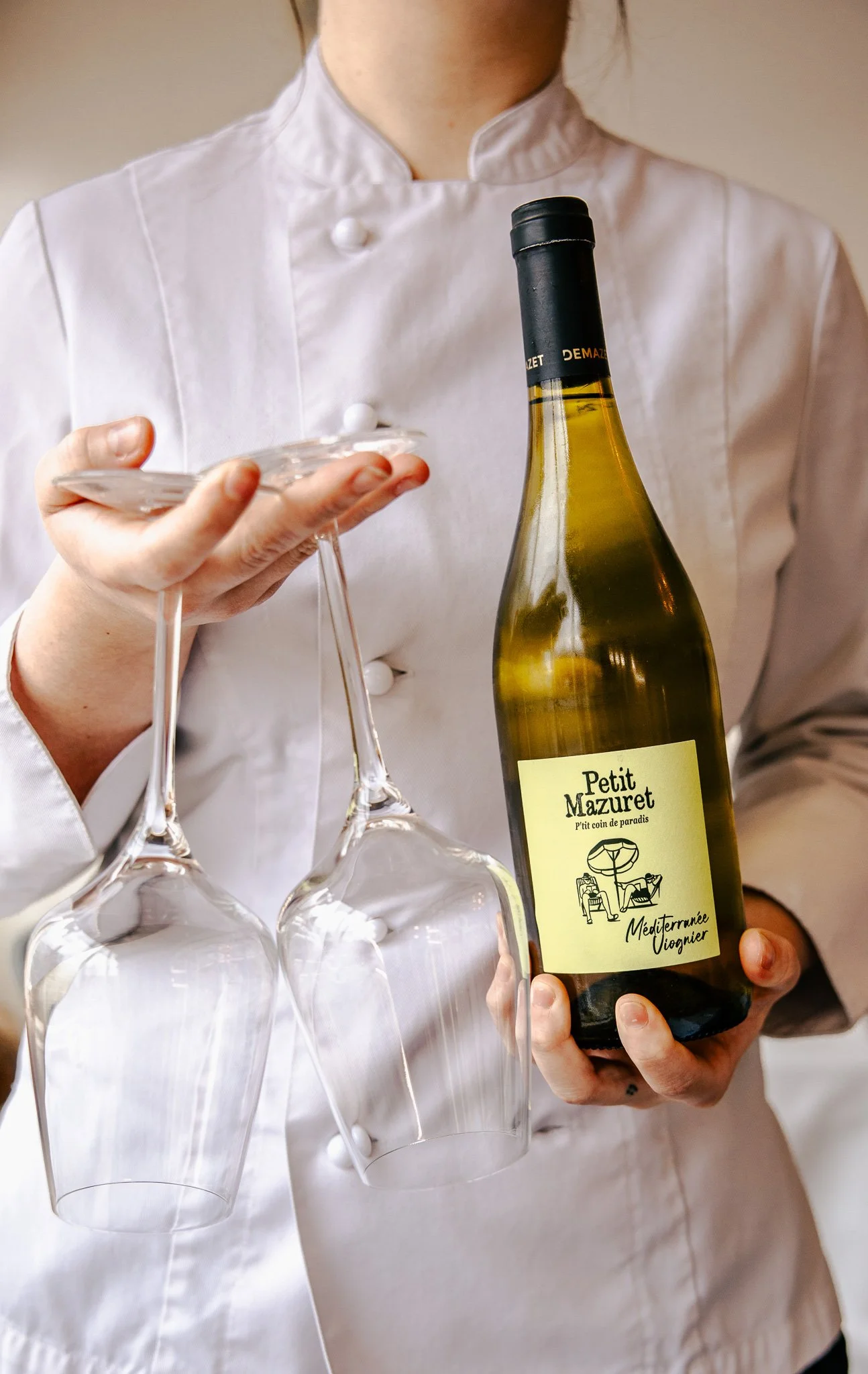 A person dressed in a white chef's coat holding a bottle of wine labeled Petit Mazuret and several empty wine glasses.