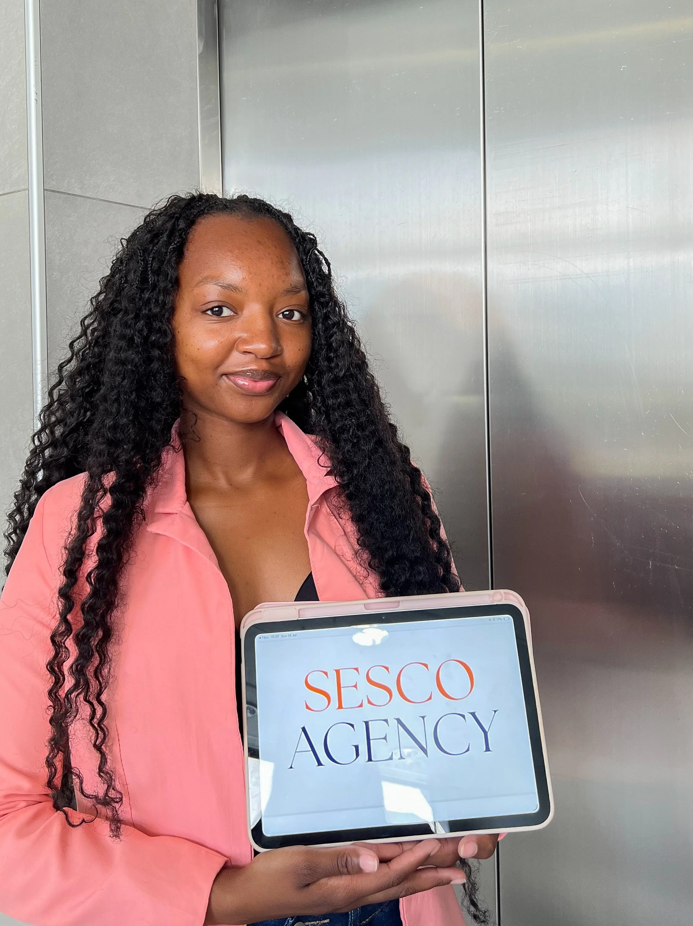 A woman with long curly hair wearing a pink shirt is holding a sign that says 'SESCO Agency' in front of an elevator.