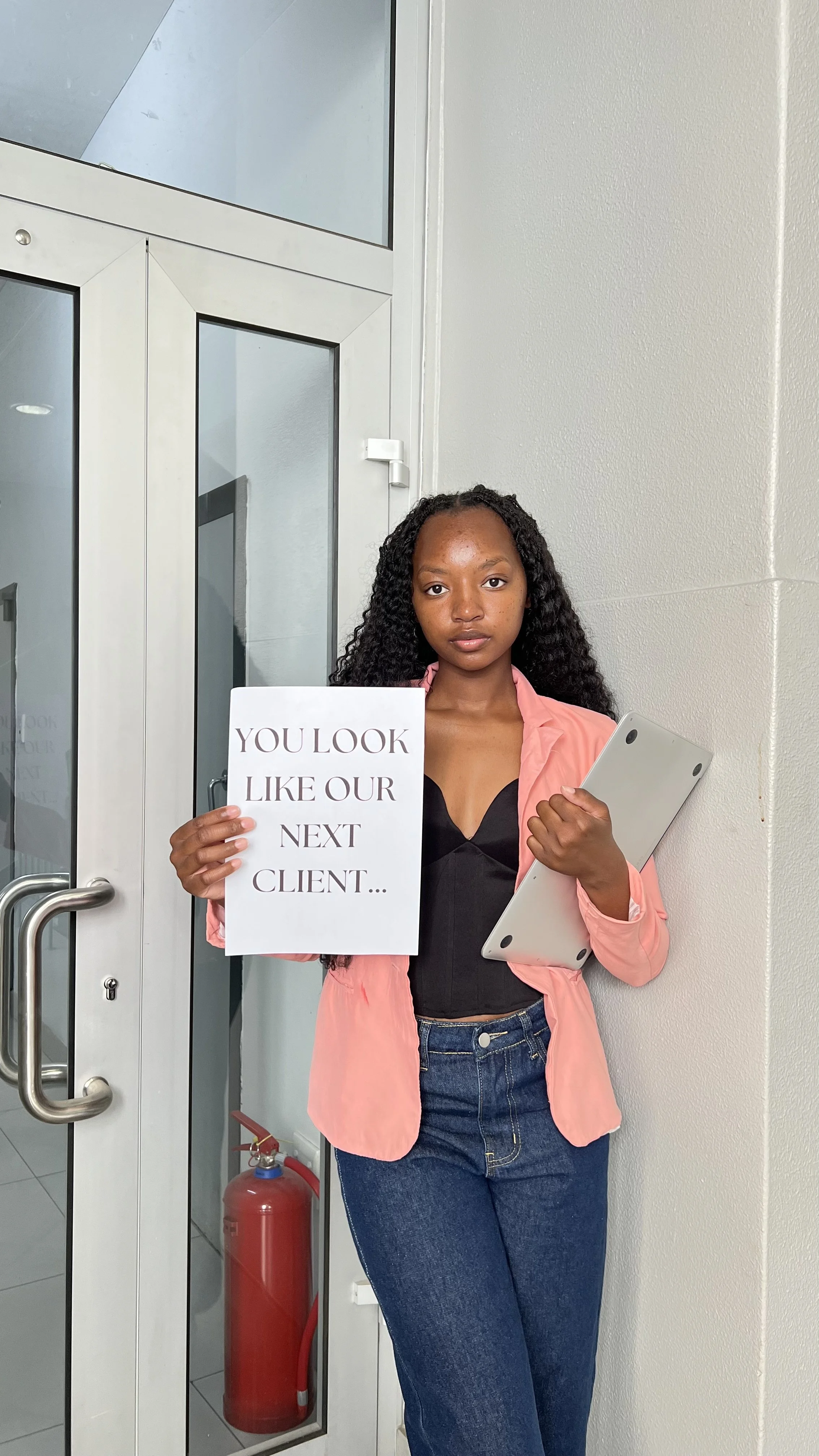 A woman standing indoors near a door, holding a sign that reads 'You look like our next client...' and a laptop, wearing a pink jacket and jeans.