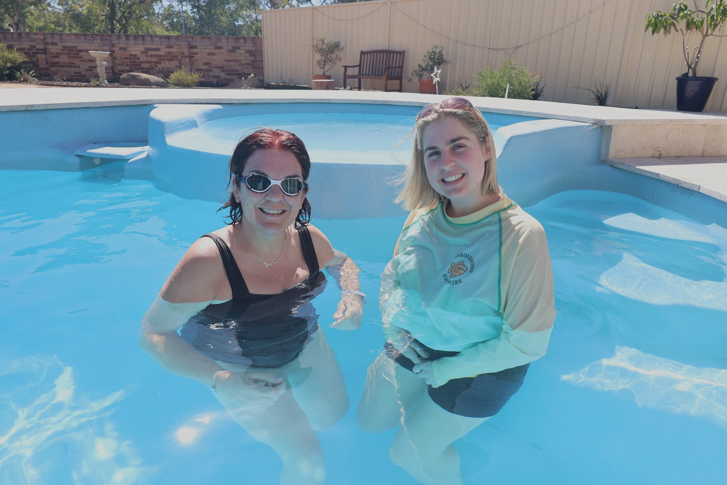 a-woman-and-a-girl-are-smiling-and-standing-in-a-swimming-pool-with-clear-blue-water-the-woman-is-wearing-a-black-swimsuit-and-sunglasses-and-the-girl-is-wearing-a-yellow-long-sleeve-shirt-the-backyard-has-potted-plants-a-wooden-bench-and-a-brick-wal
