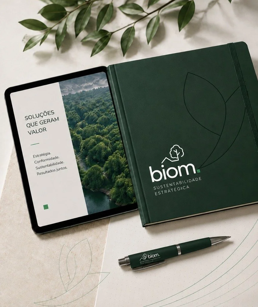 Caderno verde com o logo Biom e a frase 'Sustentabilidade Estratégica', acompanhado por uma caneta também com o logo Biom, além de folhas verdes. Também há uma tela de tablet exibindo uma apresentação sobre soluções que geram valor, estratégia, conformidade, sustentabilidade e resultados juntos, com uma imagem de uma floresta.