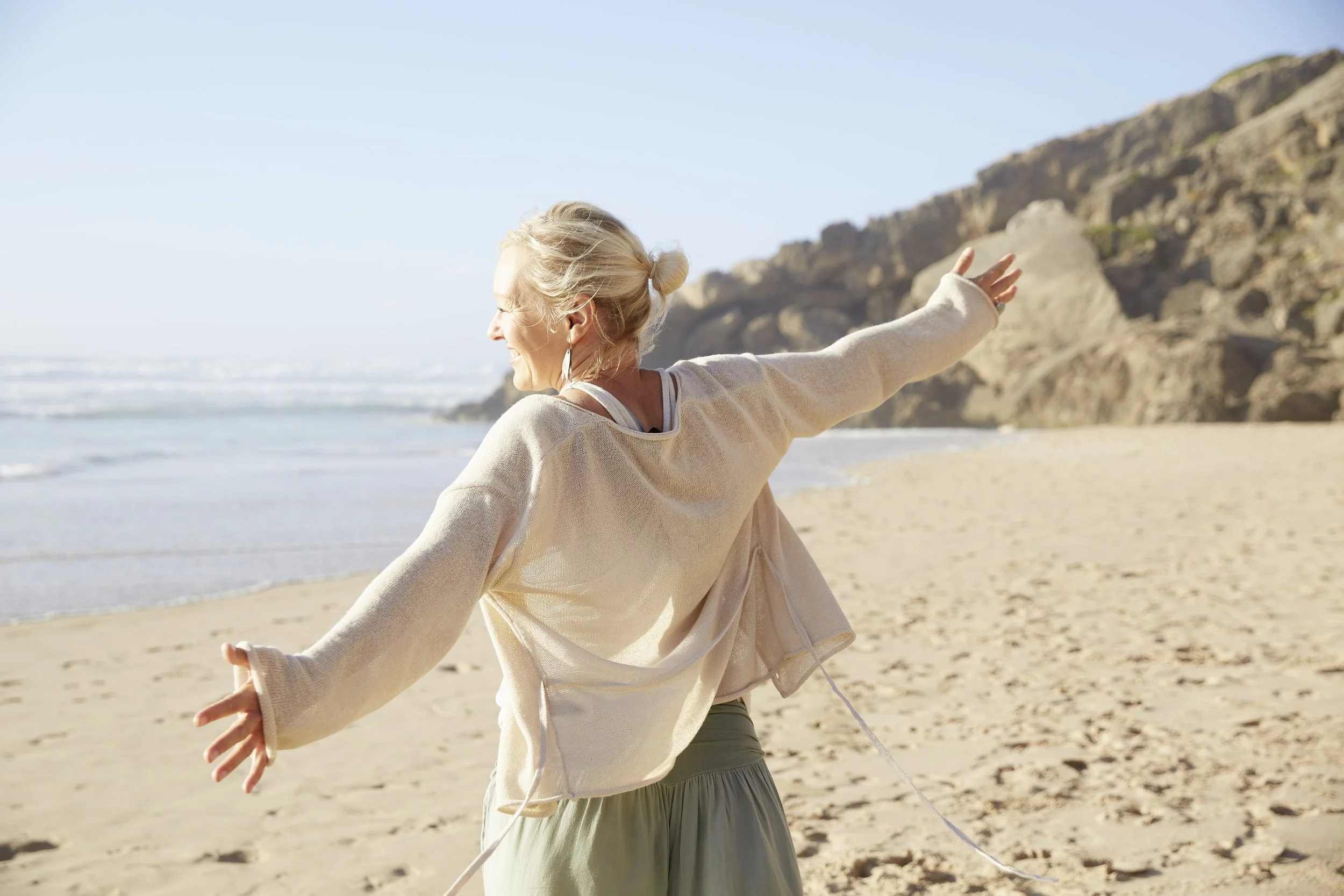A woman dressed in light-colored clothing practicing yoga or meditation on a sandy beach with rocky cliffs in the background, enjoying a sunny day by the ocean.
